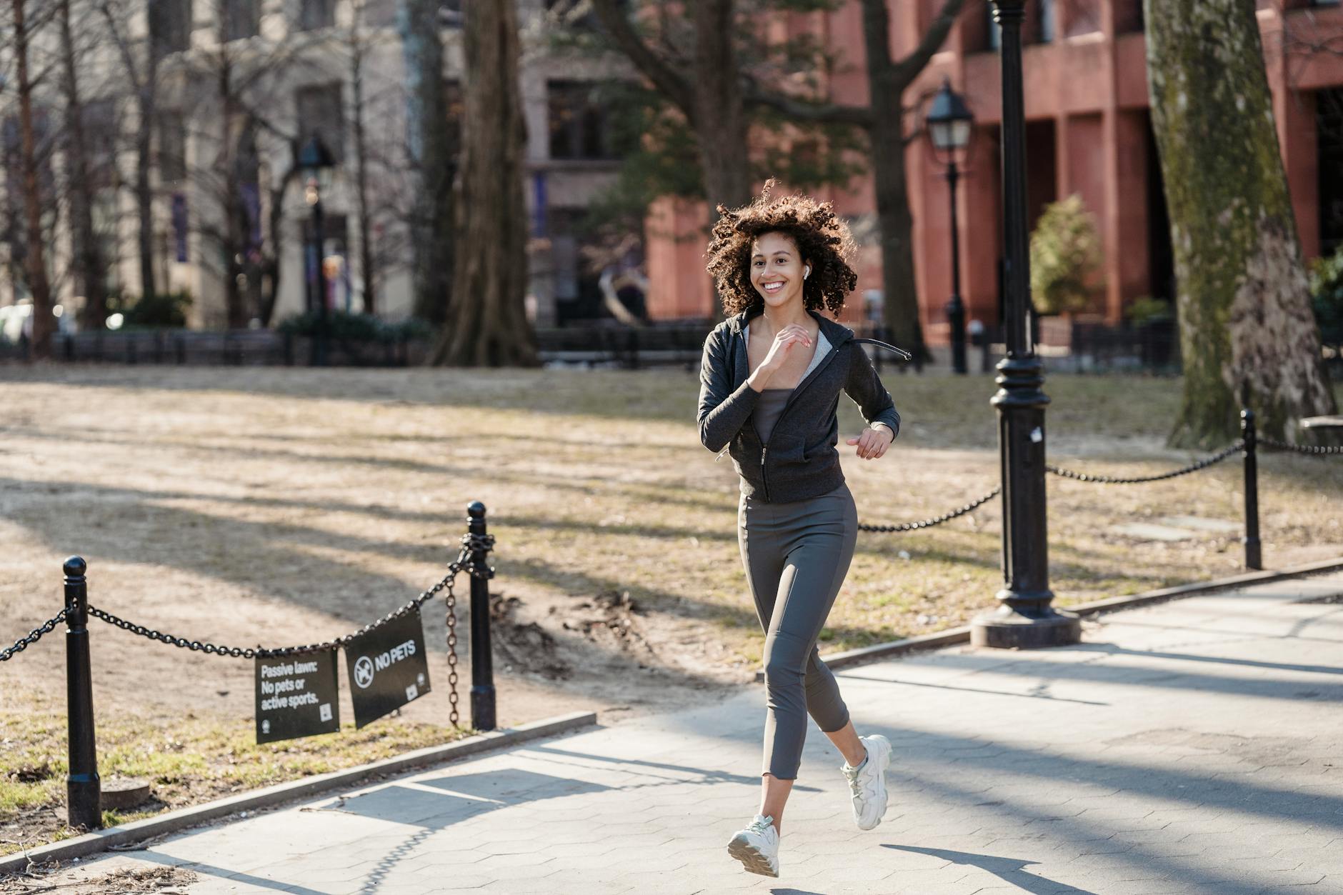 Full body of African American female jogger in sportswear smiling and running in park - spring cardio outdoors