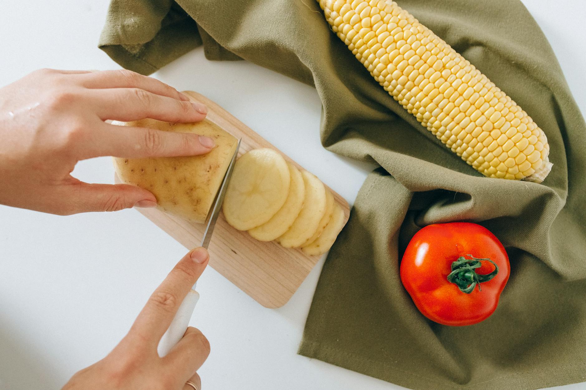 A detailed shot of hands slicing potatoes with corn and tomato on a kitchen counter. - spring meal prep
