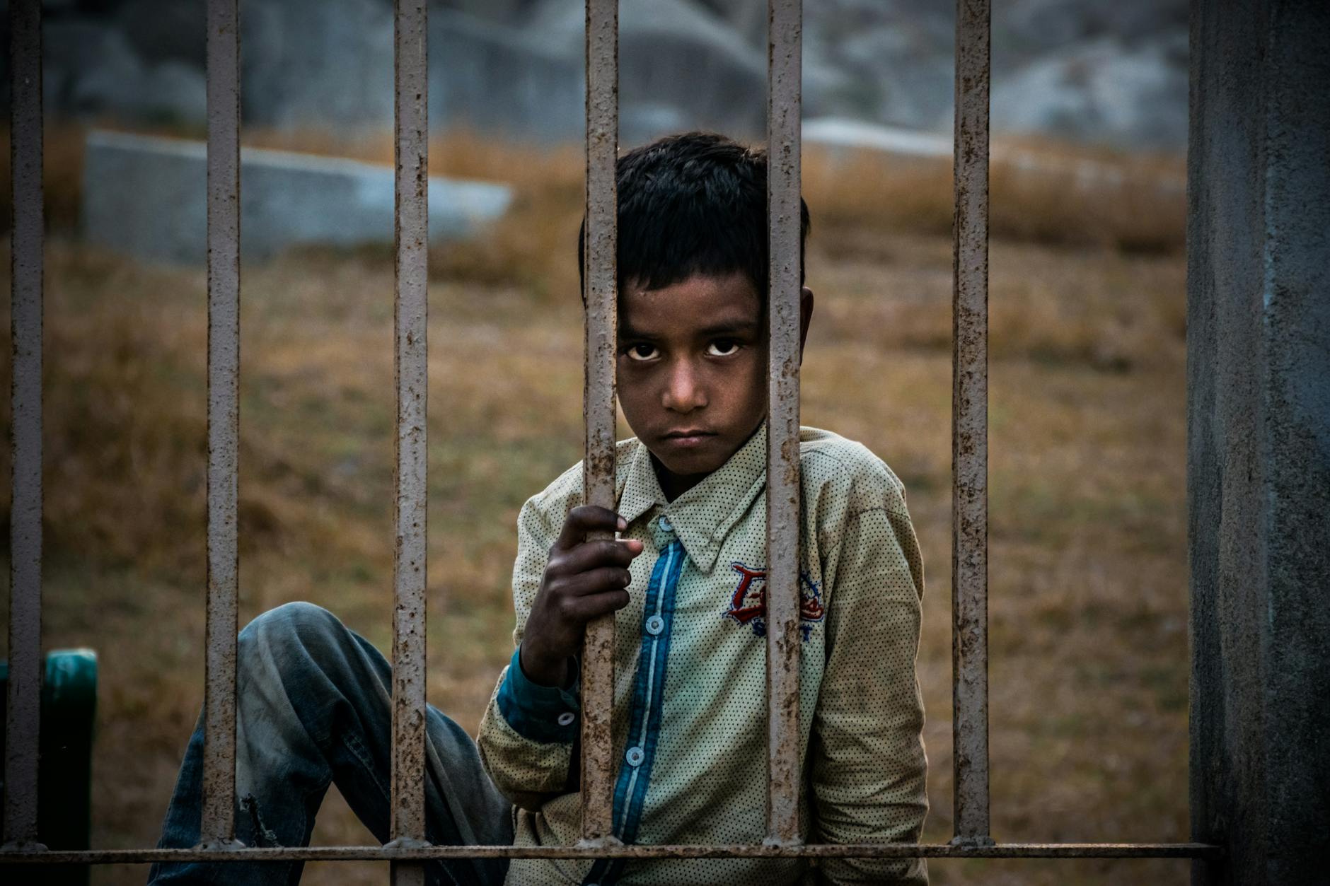 A boy with a serious expression sits behind a rusted fence outdoors, conveying a sense of contemplation. - spring sad science