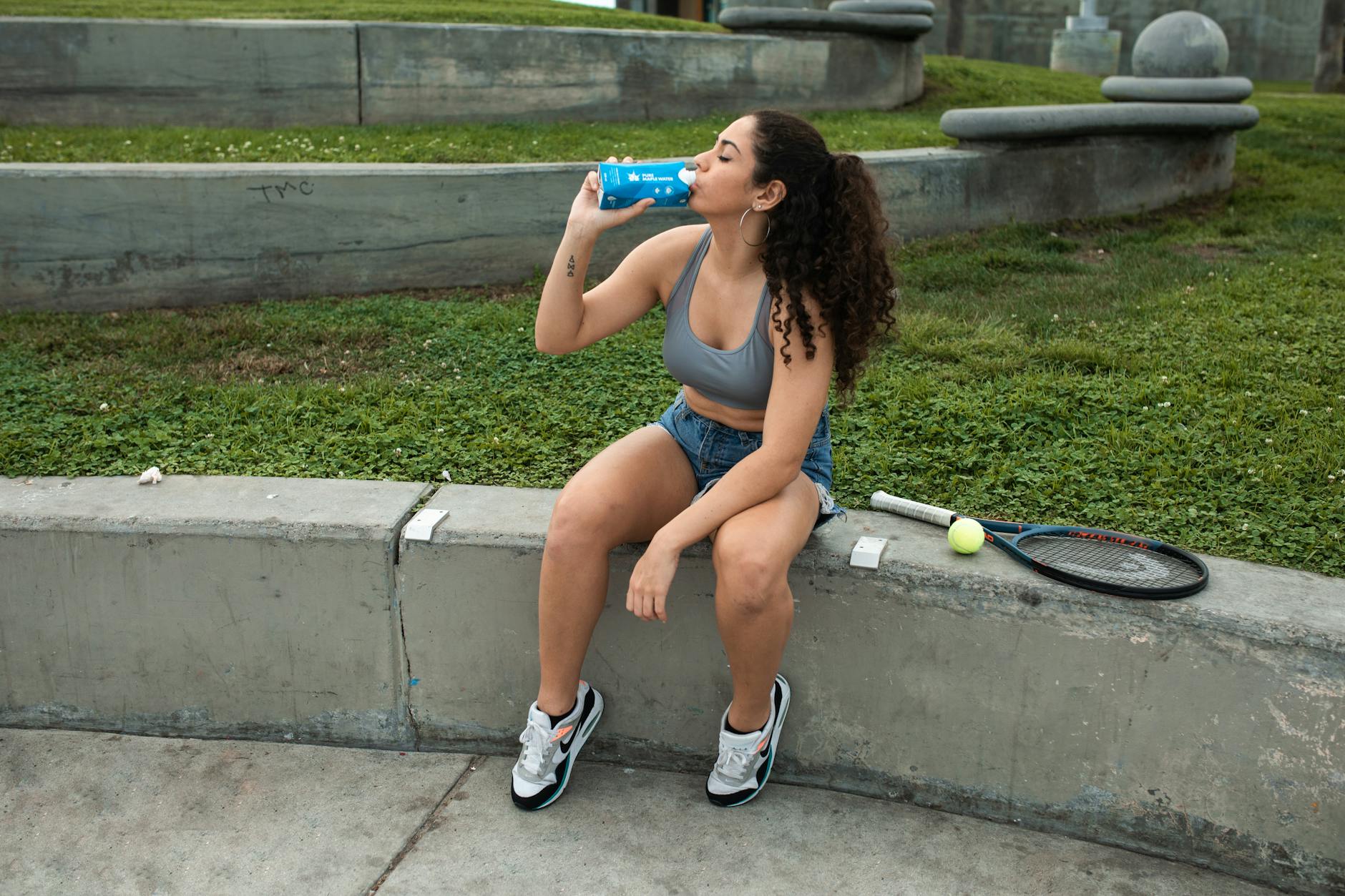 Young woman sitting on a bench with tennis racket and drinking water in a park. - spring workout refresh