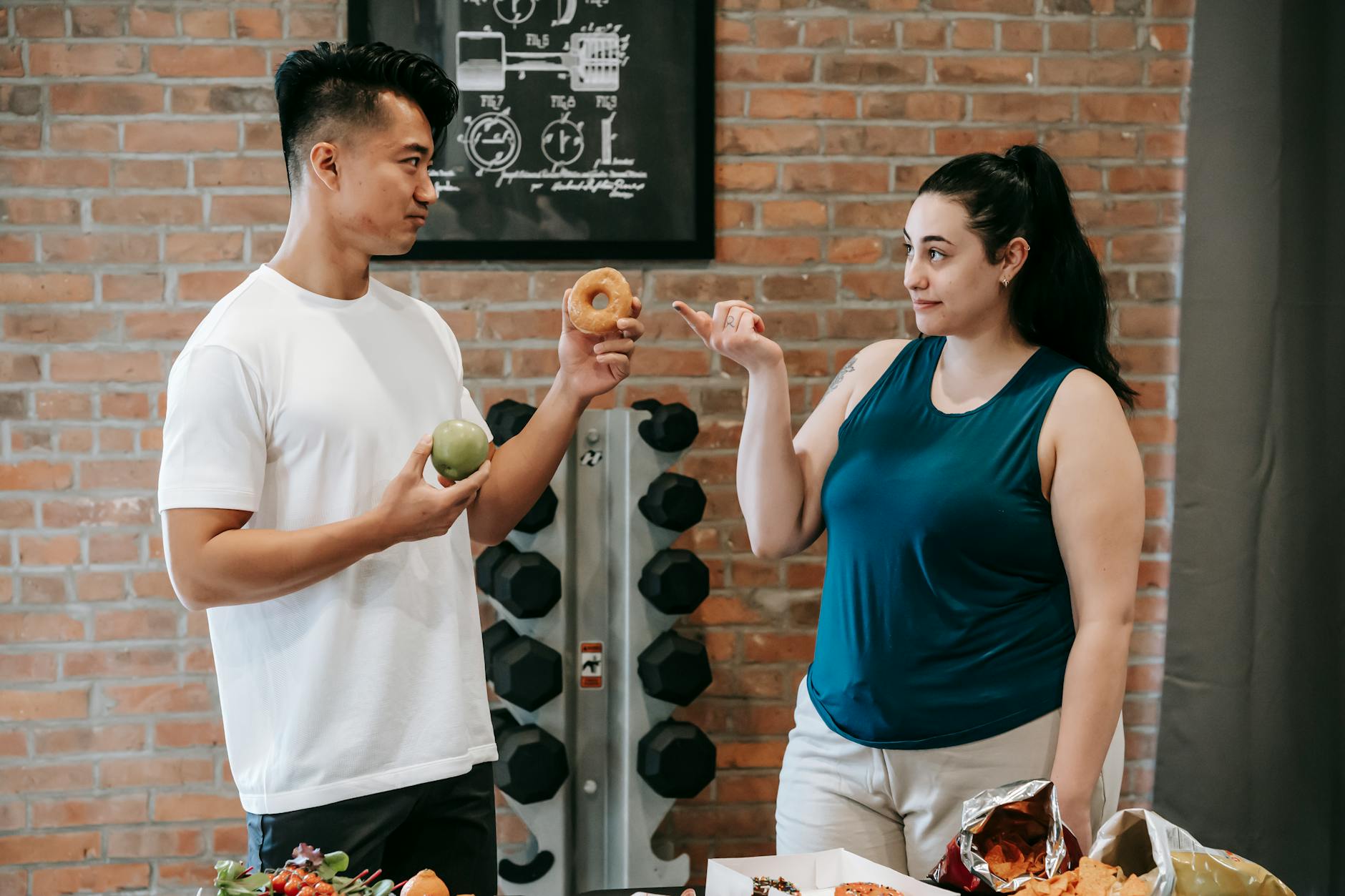 Side view of Asian personal instructor with apple and doughnut offering healthy food to plus size woman while standing in gym - teen weight loss