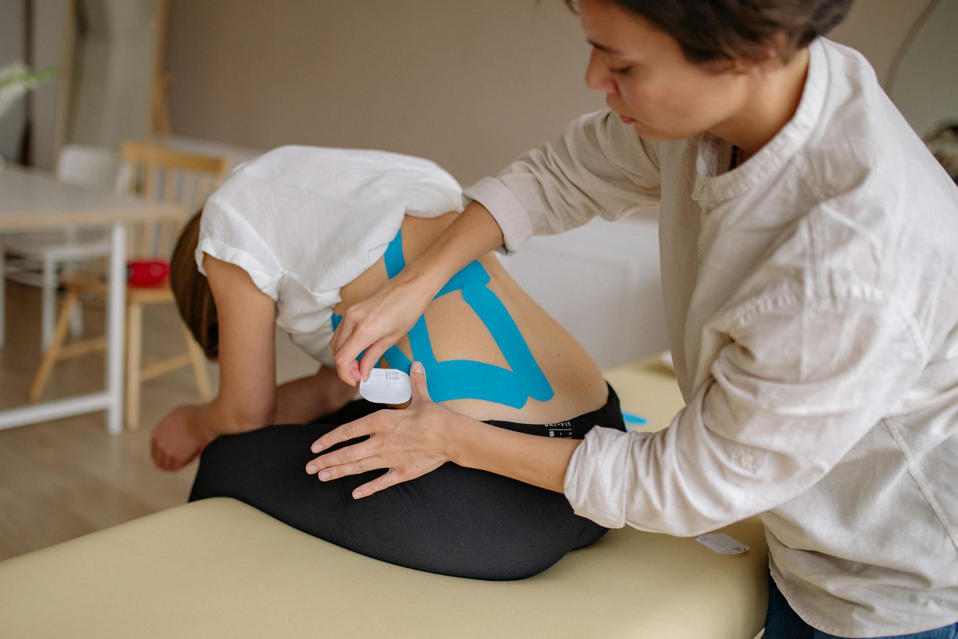 A therapist applies blue kinesiology tape to a patient's back in a clinical setting. - back pain relief