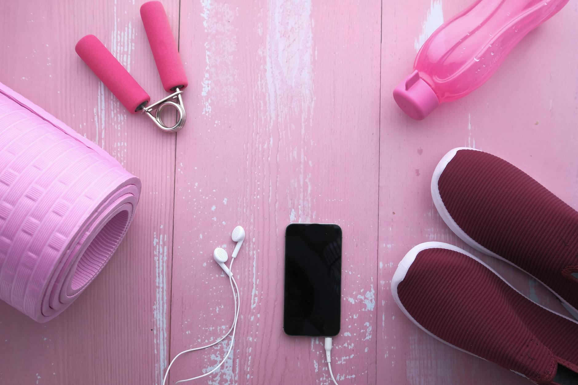 Top view of fitness accessories including yoga mat, shoes, water bottle, and smartphone on pink wood. - beginner workouts