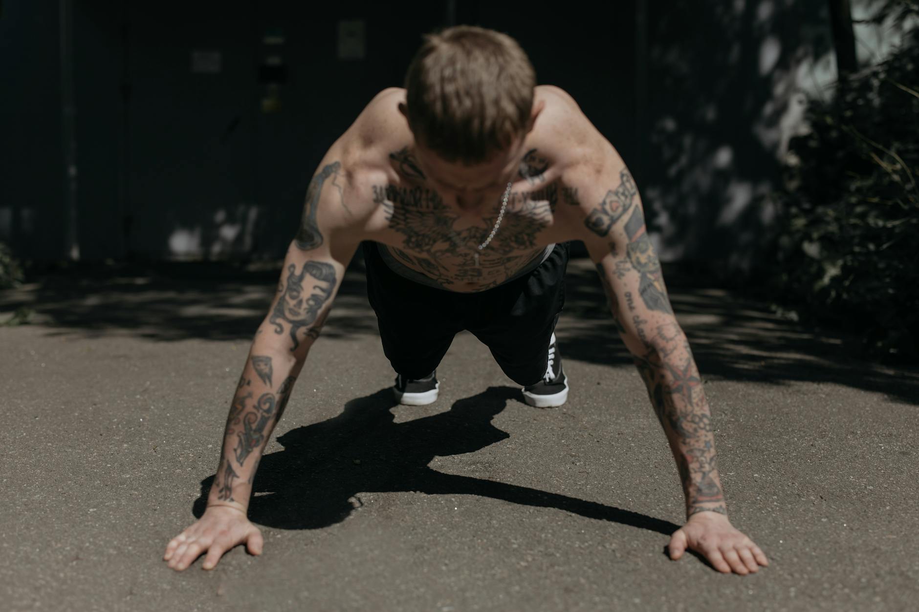 Shirtless tattooed man performing push-ups on a concrete path outdoors, showcasing fitness and endurance. - bodyweight strength reset