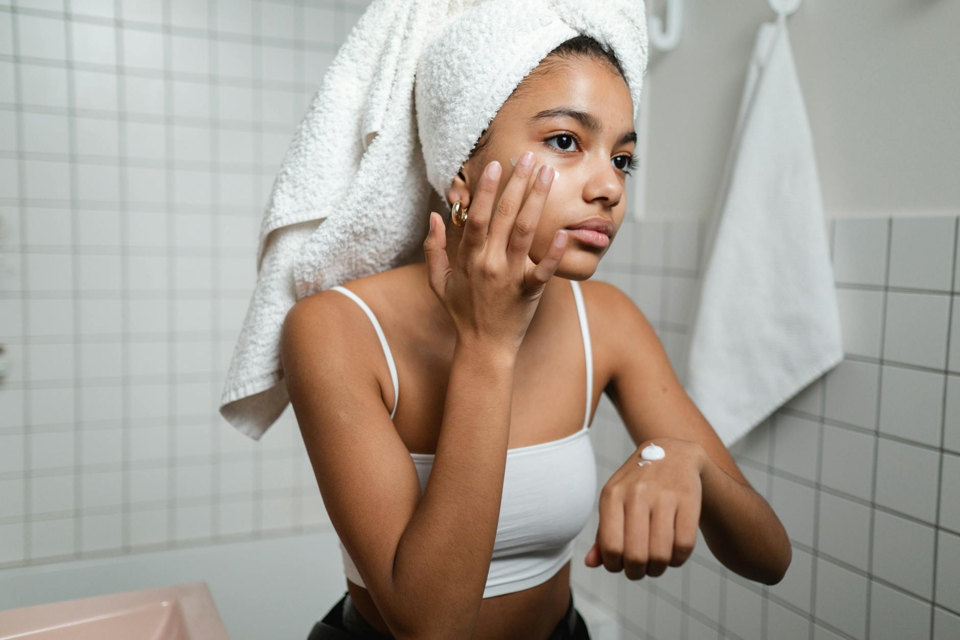 A young woman in a bathroom applying skincare cream with a towel on her head, focusing on self-care. - daily skincare routine