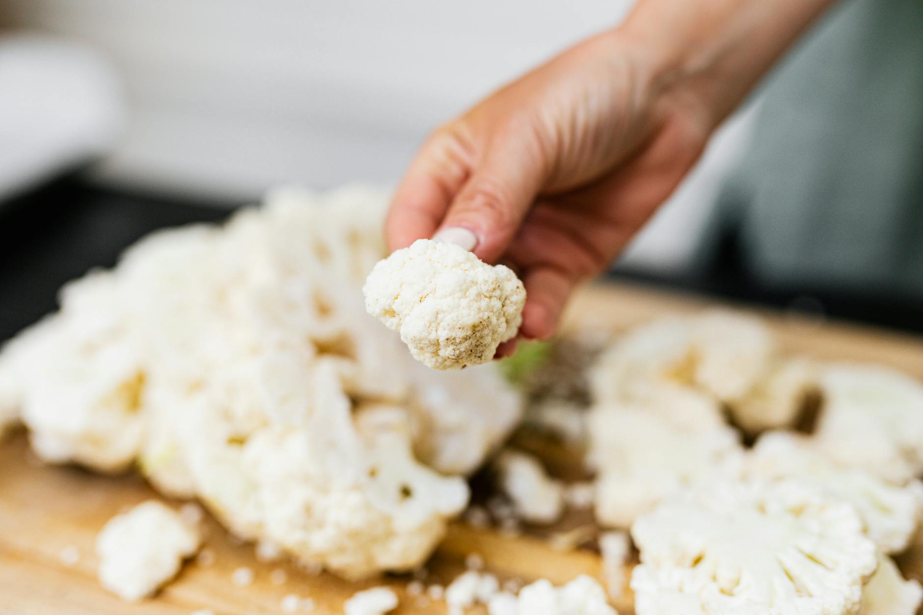 A hand holds a fresh cauliflower floret over a wooden board with scattered florets, showcasing a cooking preparation. - easy low carb dinners