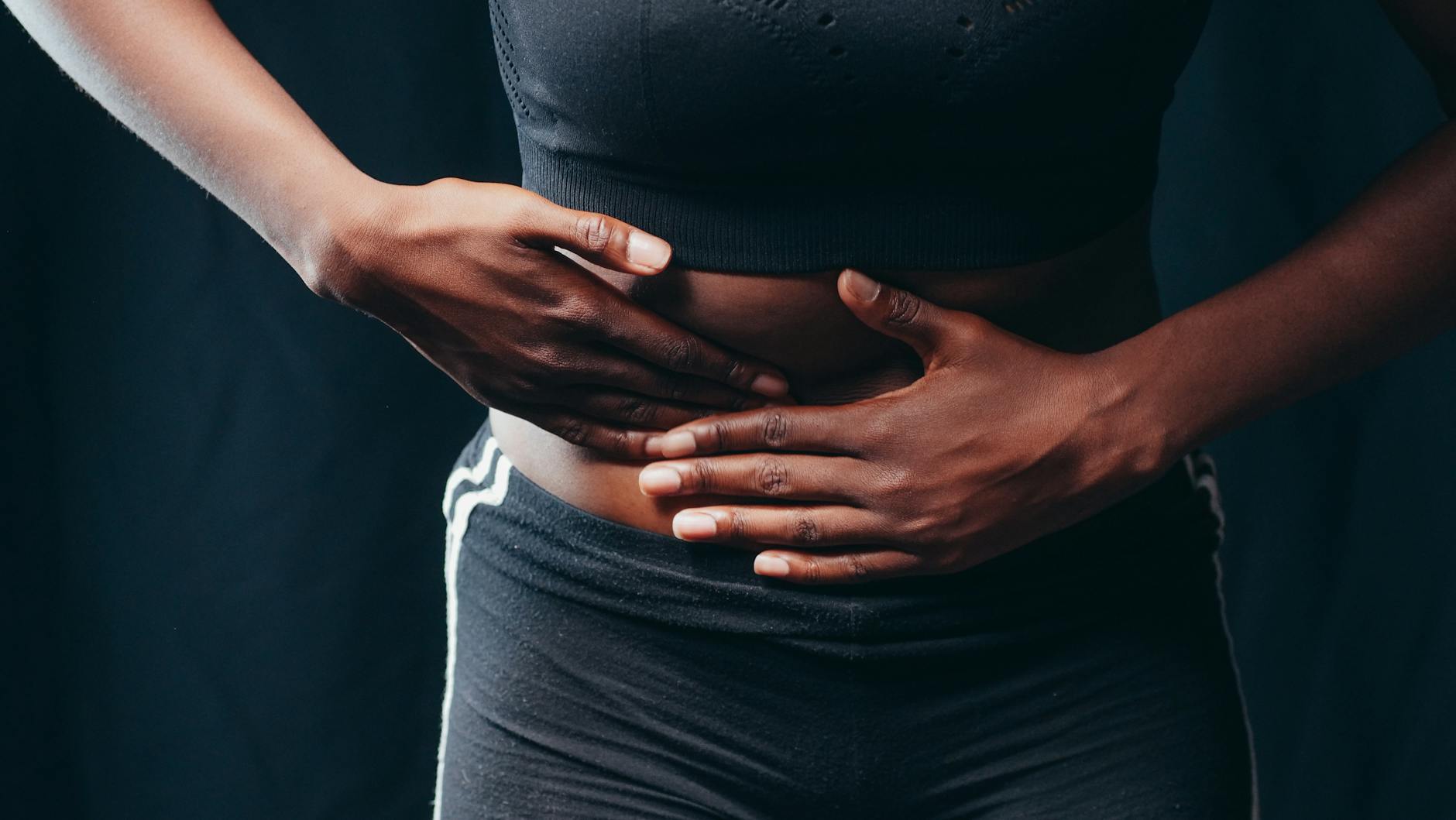 Close-up of a person pressing hands on abdomen, indicating discomfort or pain, against a black background. - gut health reset
