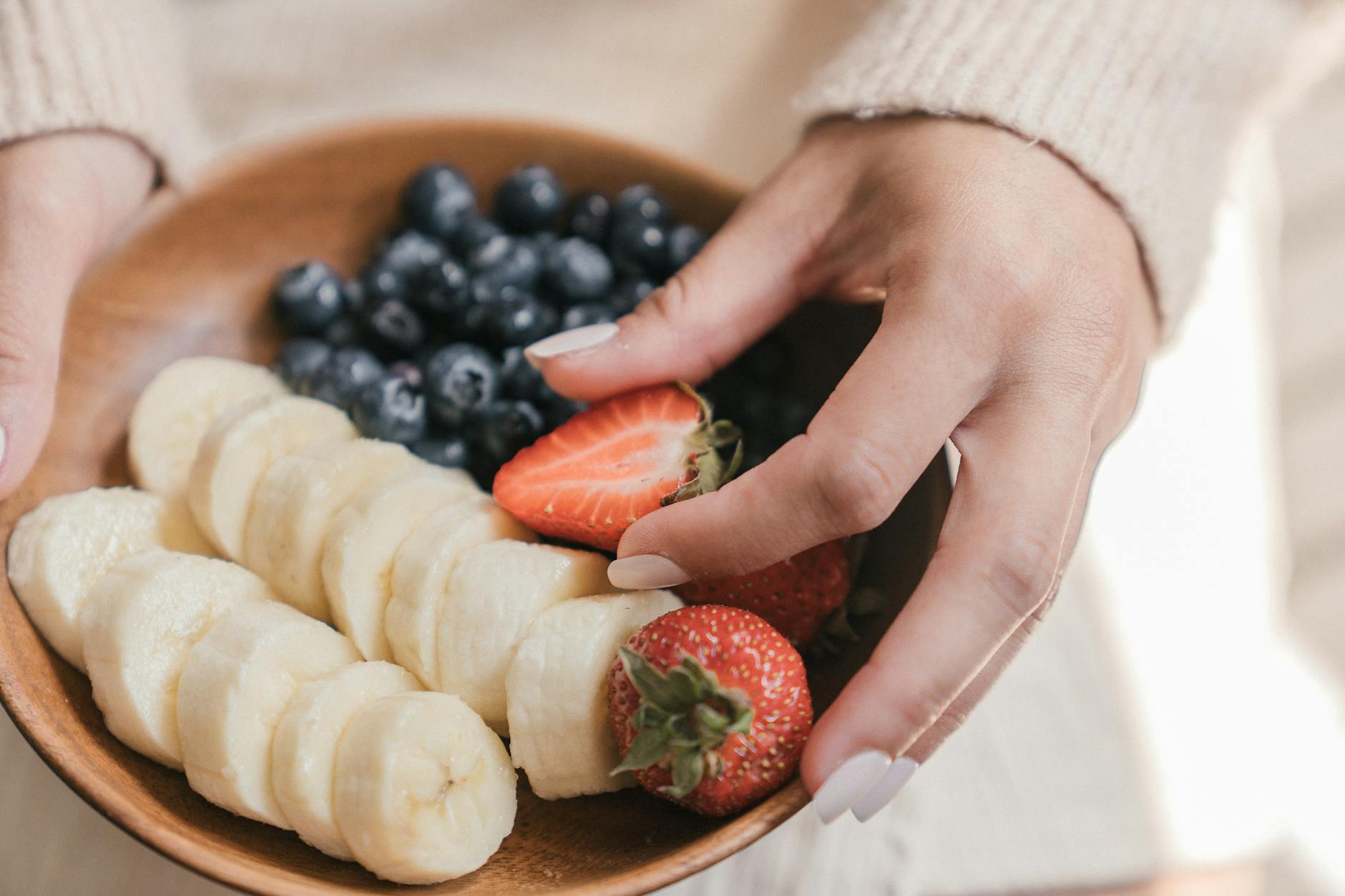 A wooden bowl filled with fresh bananas and berries held by an unrecognizable person's hands. - healthy spring snacks