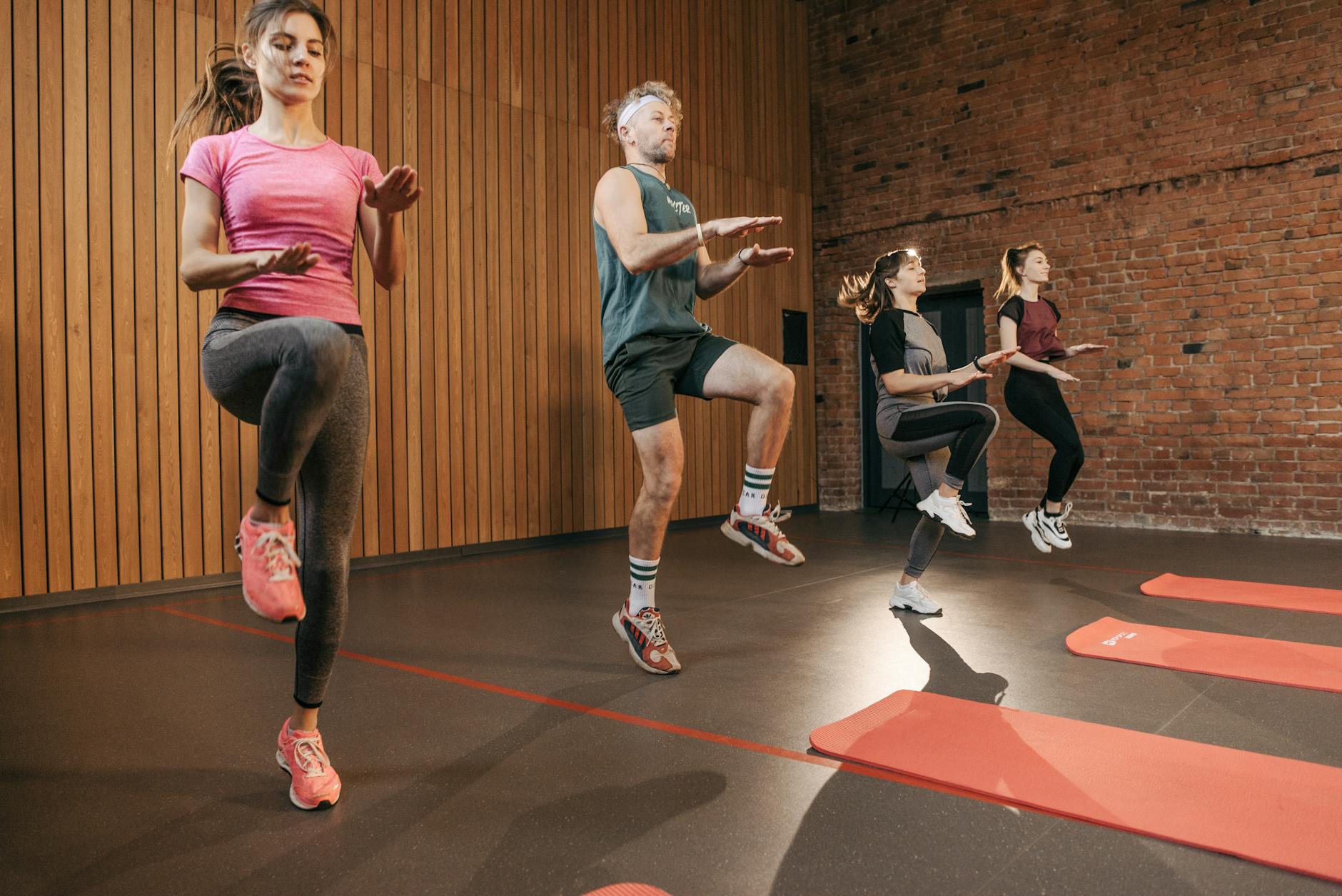 Four adults performing high knees in an indoor gym with wooden panel walls. - interval training cardio