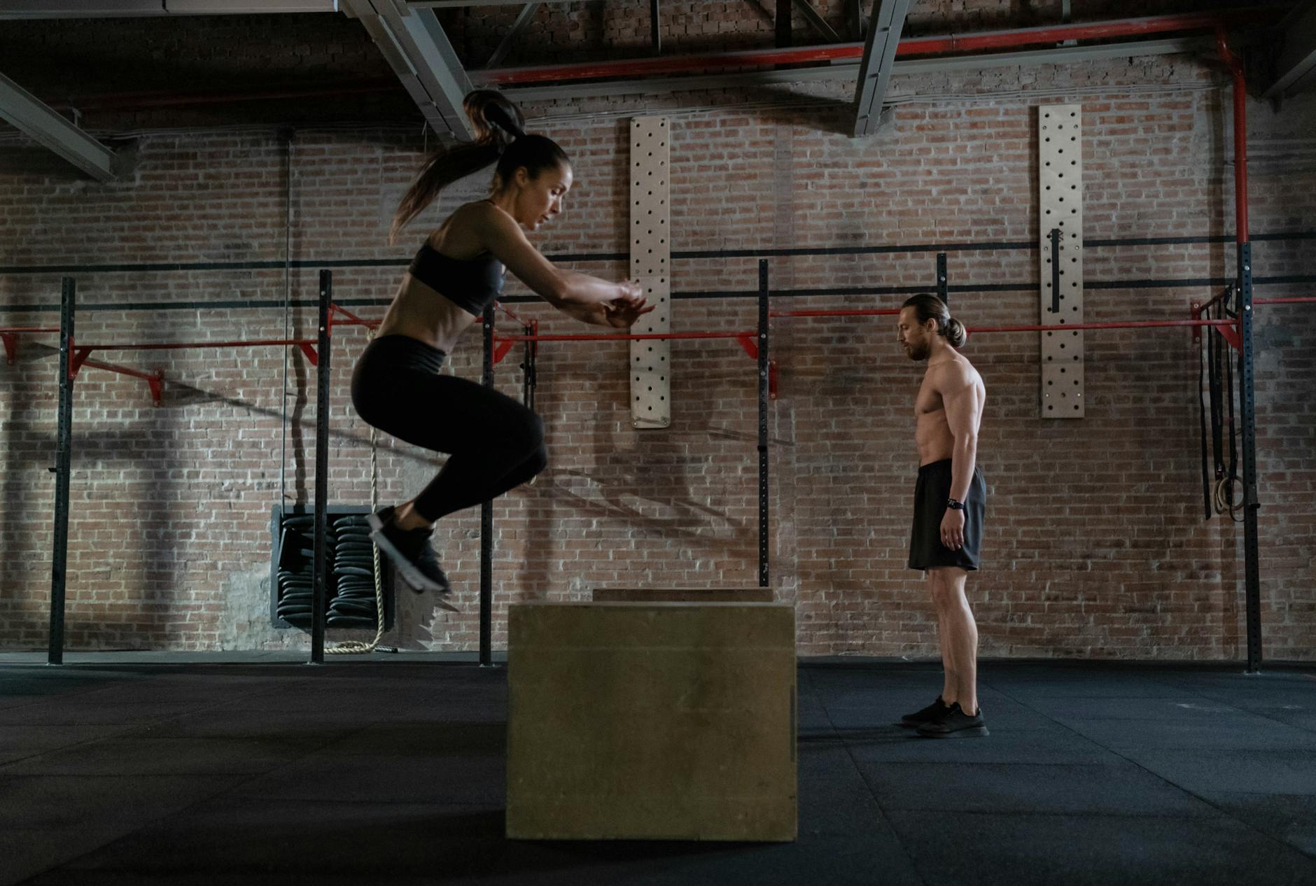 Female athlete performing box jump in gym watched by coach. Intense fitness training indoors. - interval training fitness