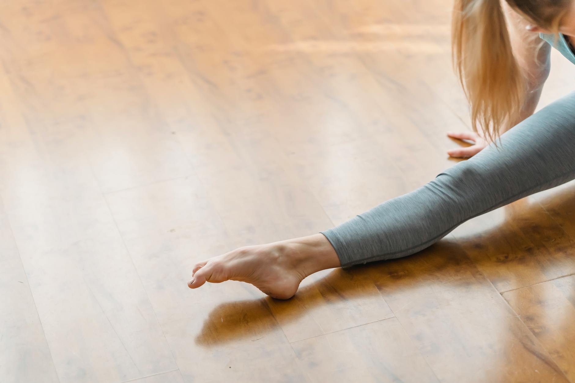 Crop anonymous barefoot female doing splits on wooden floor in bright daylight at home - lower body flexibility