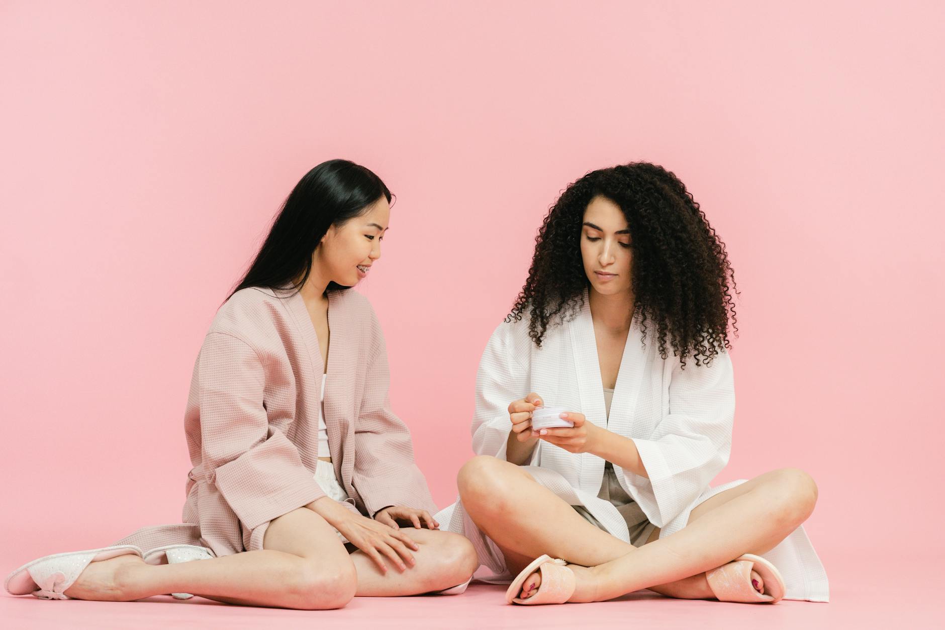 Two women in robes discussing skincare cream in a calm, pink setting. - magnesium for women