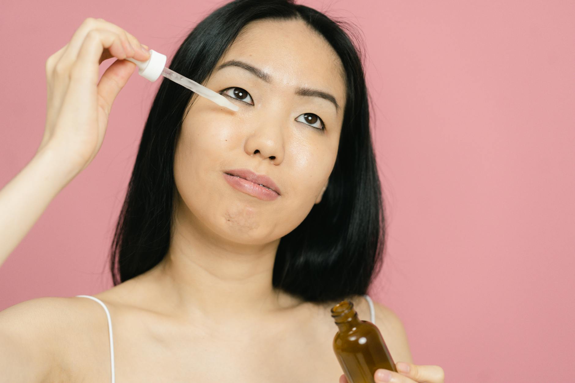 A woman applying facial serum with a dropper for skin care on a pink background. - natural anti aging skin