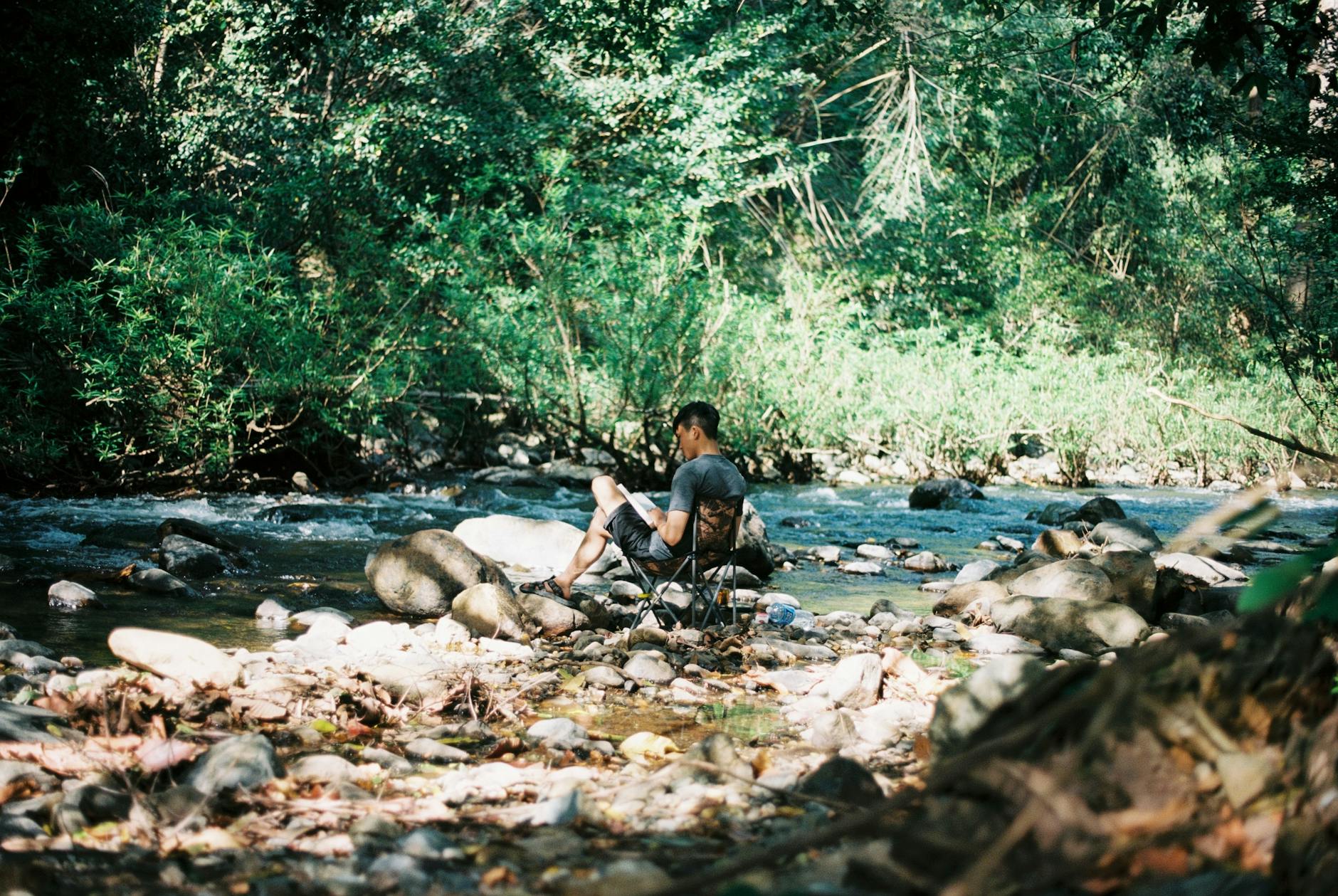 A man sits on a chair by a tranquil stream in a lush forest, enjoying a peaceful moment. - nature digital detox
