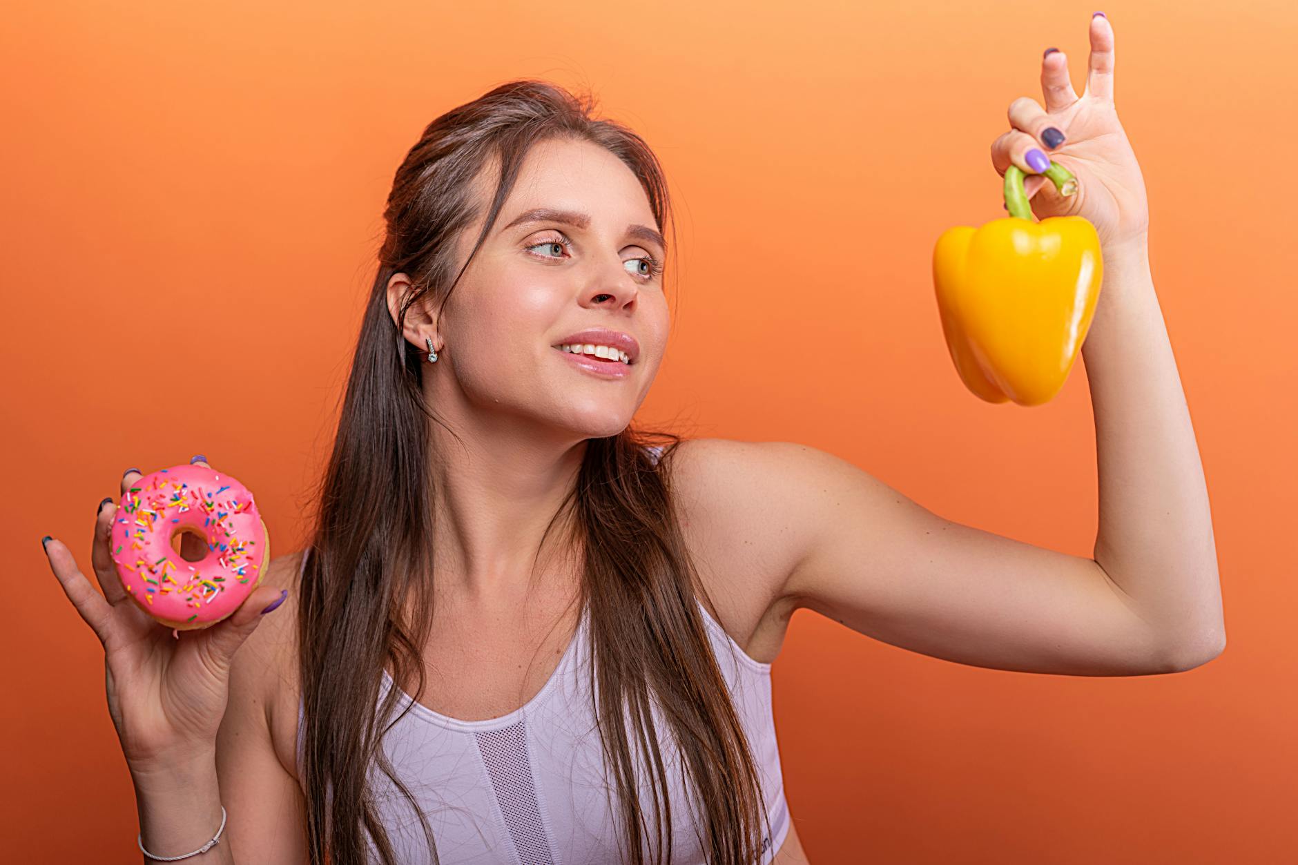 Woman in sports bra juggling choice between a donut and bell pepper on orange background. - optimal nutrition guide