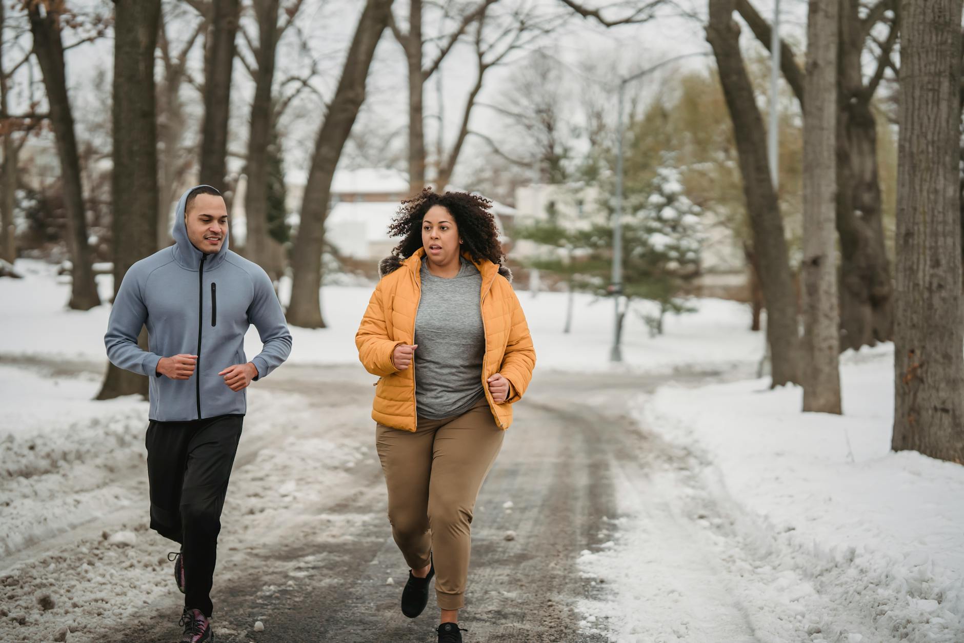 Full body of plus size African American female in warm clothes running with black coach on snowy pathway in park during training on winter time - post winter workout