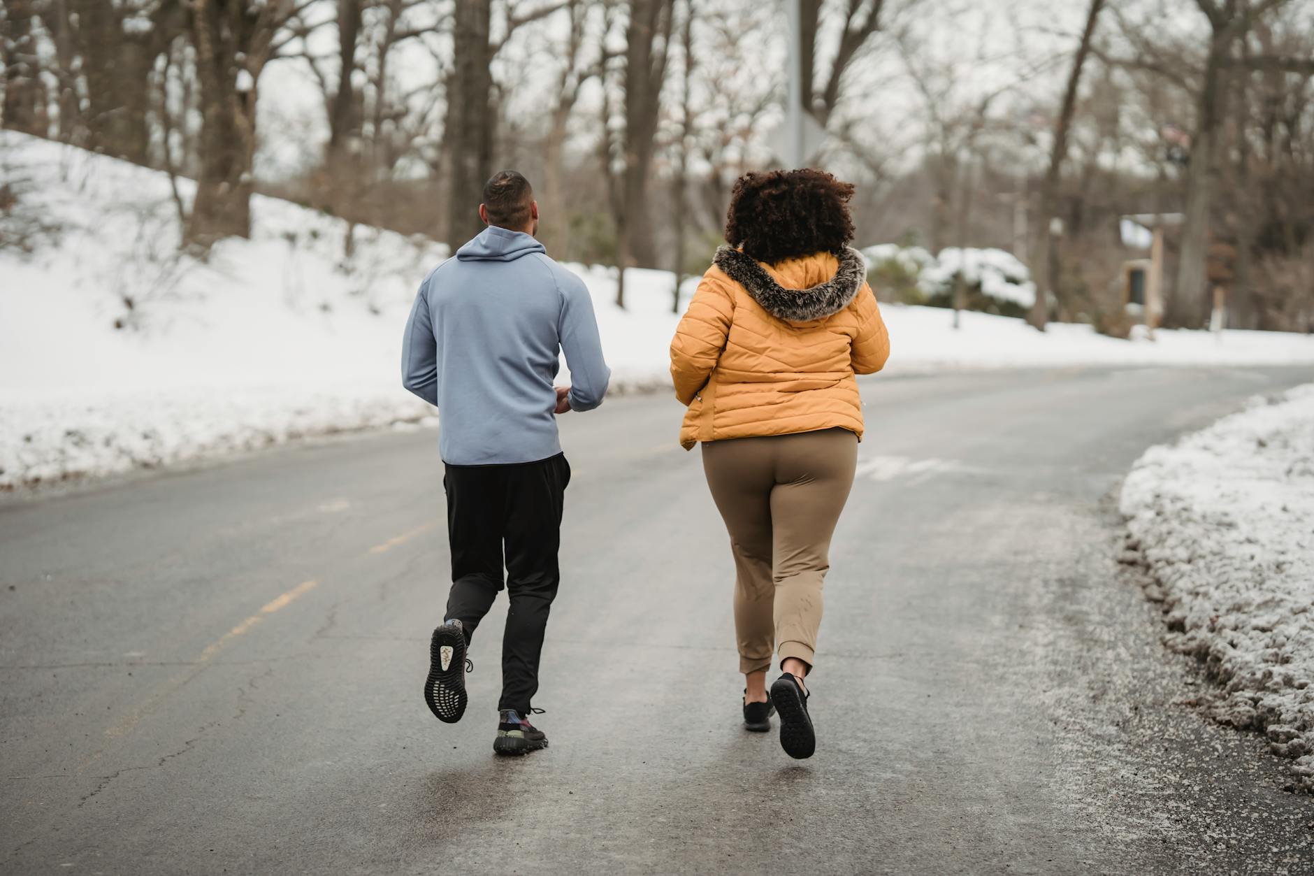 Back view full length fit male fitness trainer and plump female in warm jacket jogging together on asphalt roadway in snowy forested suburb on cold winter weather - post winter workouts