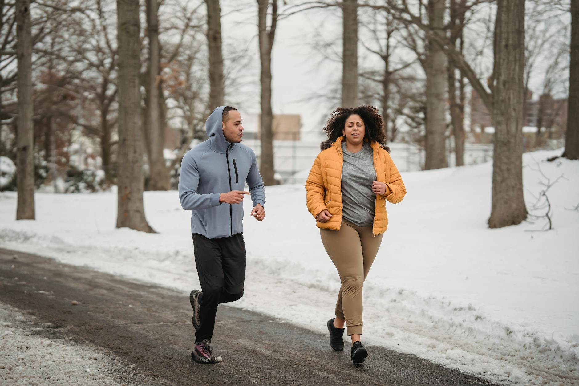 Full body of African American male instructor running with black overweight woman on pathway among snowy ground with trees during winter time - post winter workouts
