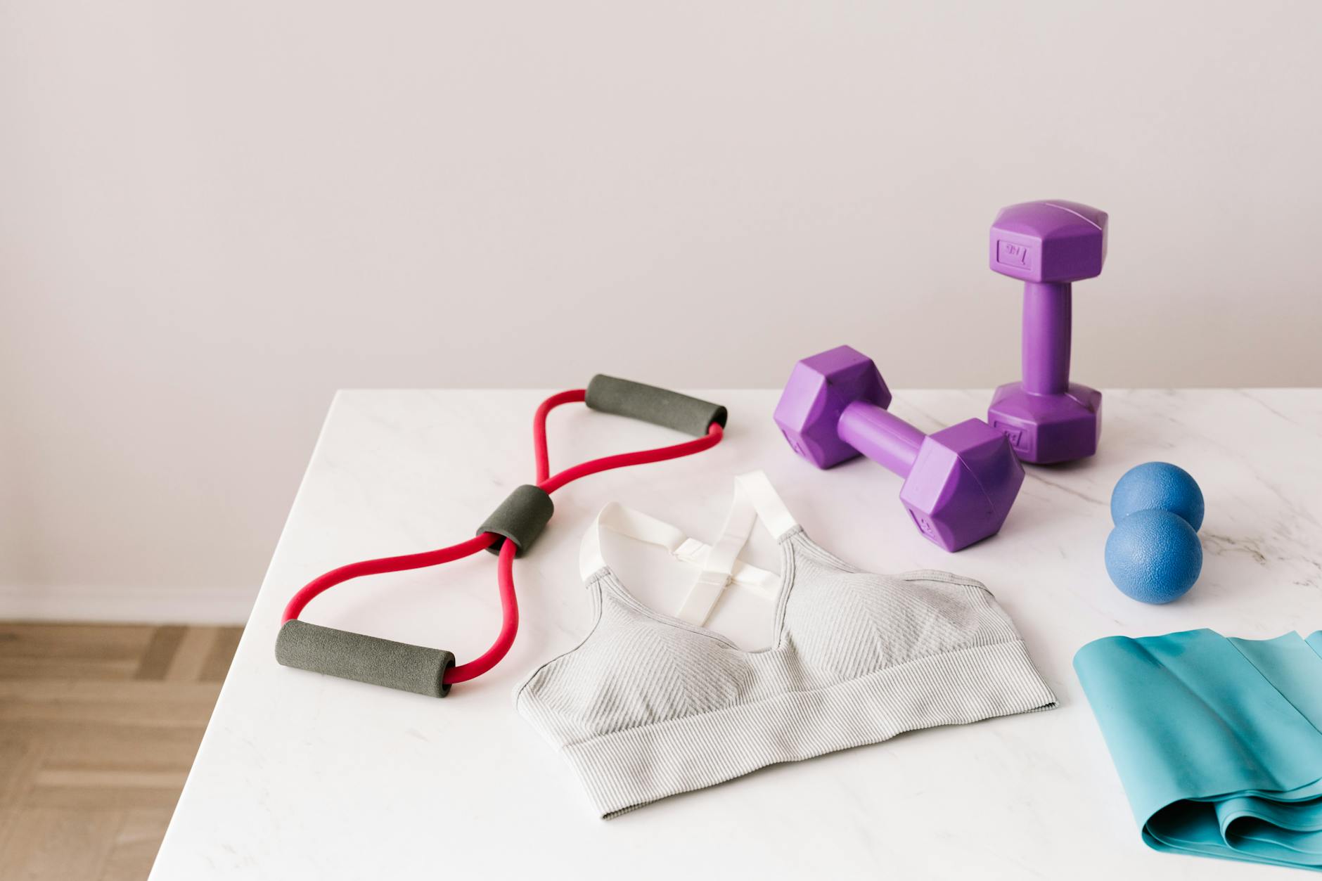 From above of white sports bra surrounded by purple dumbbells and blue tape expander and red tubular expander and blue massage double ball placed on marble surface of table - resistance training benefits