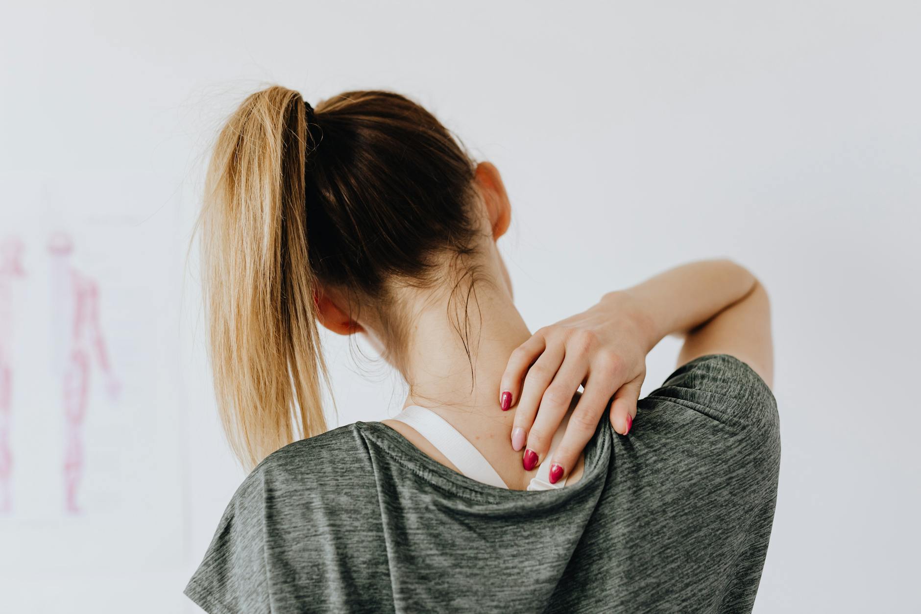 Back view of slim blond female in casual wear with manicure indicating pain spot on back while sitting in front of white wall with paper drawing in clinic - shoulder pain relief