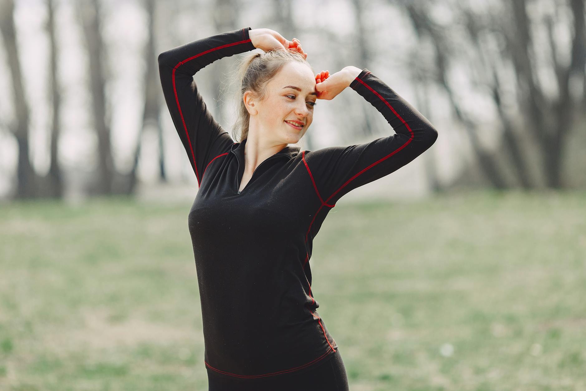 Energy young female in black sportswear standing in park and stretching arms during outdoors workout while smiling and looking away - spring allergies exercise
