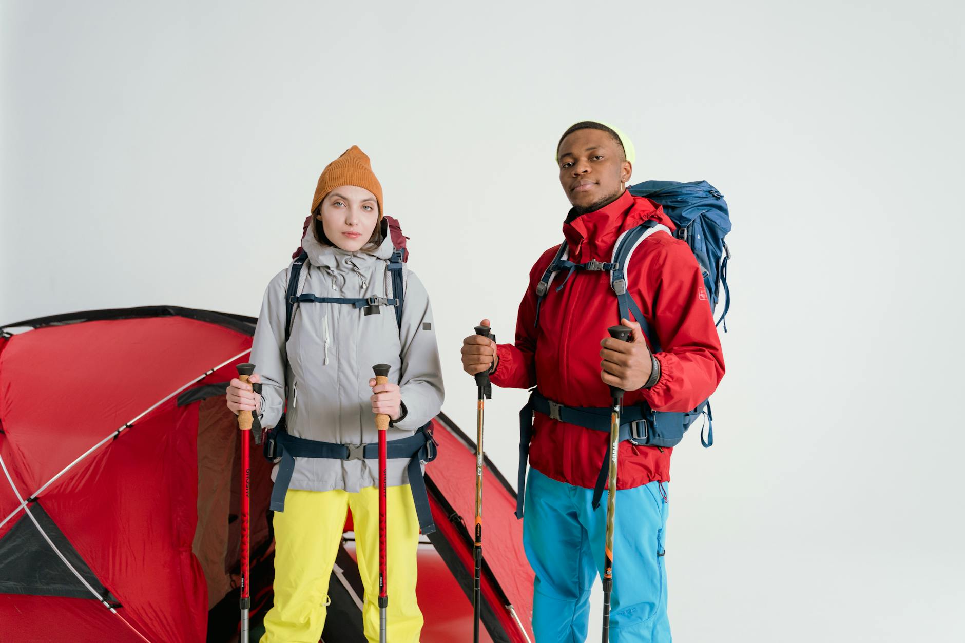 Two adults equipped with camping gear and hiking sticks pose in front of a tent on a white background. - spring hiking gear