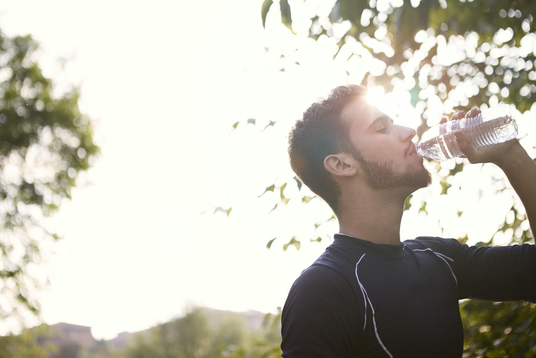 Side view of a young man drinking water from a plastic bottle outdoors in sunlight. - spring hydration plan