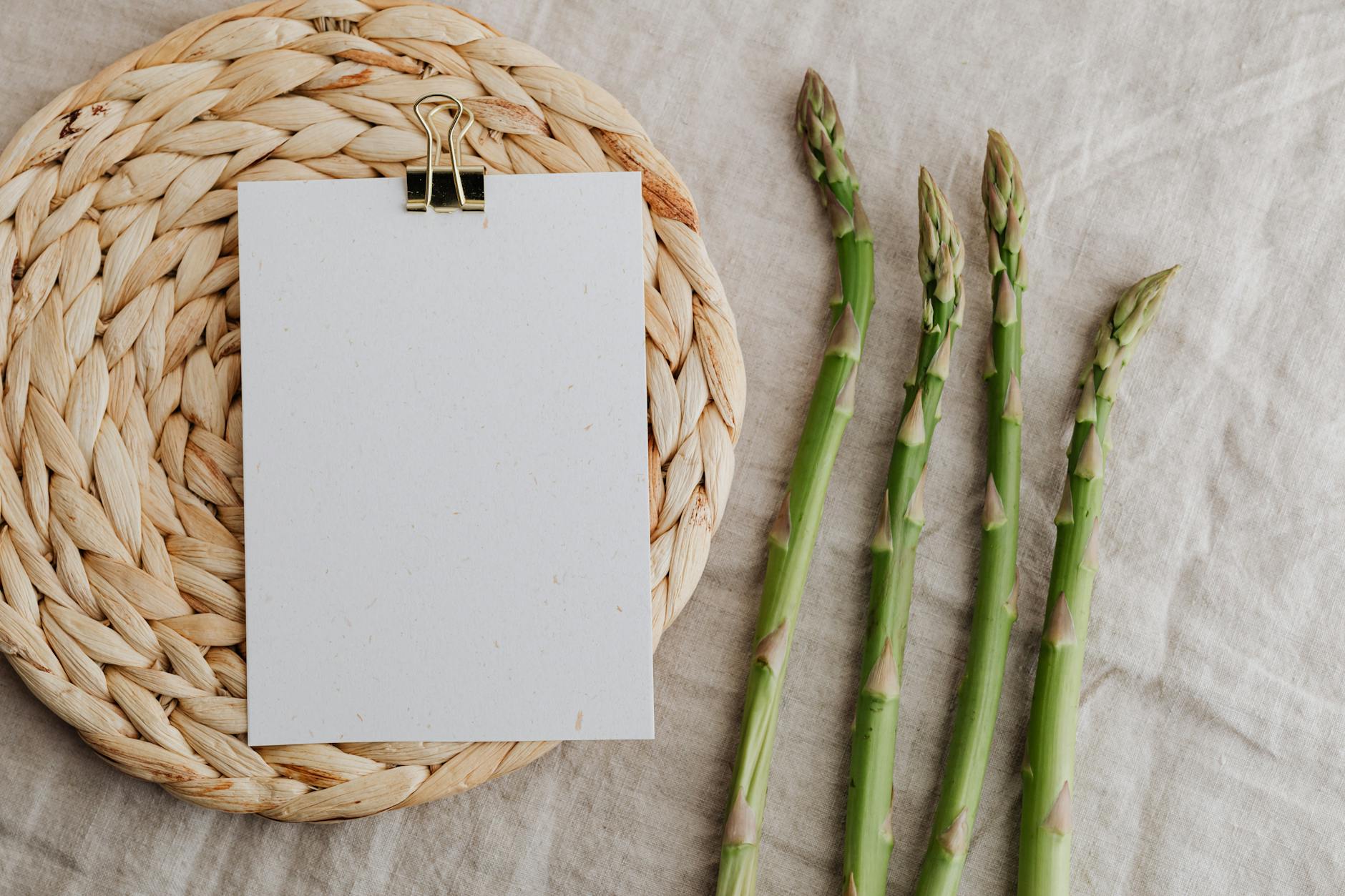 From above of four fresh green asparagus sprouts and blank sheet of paper over round wicker placemat laid on white tablecloth - spring inflammation diet