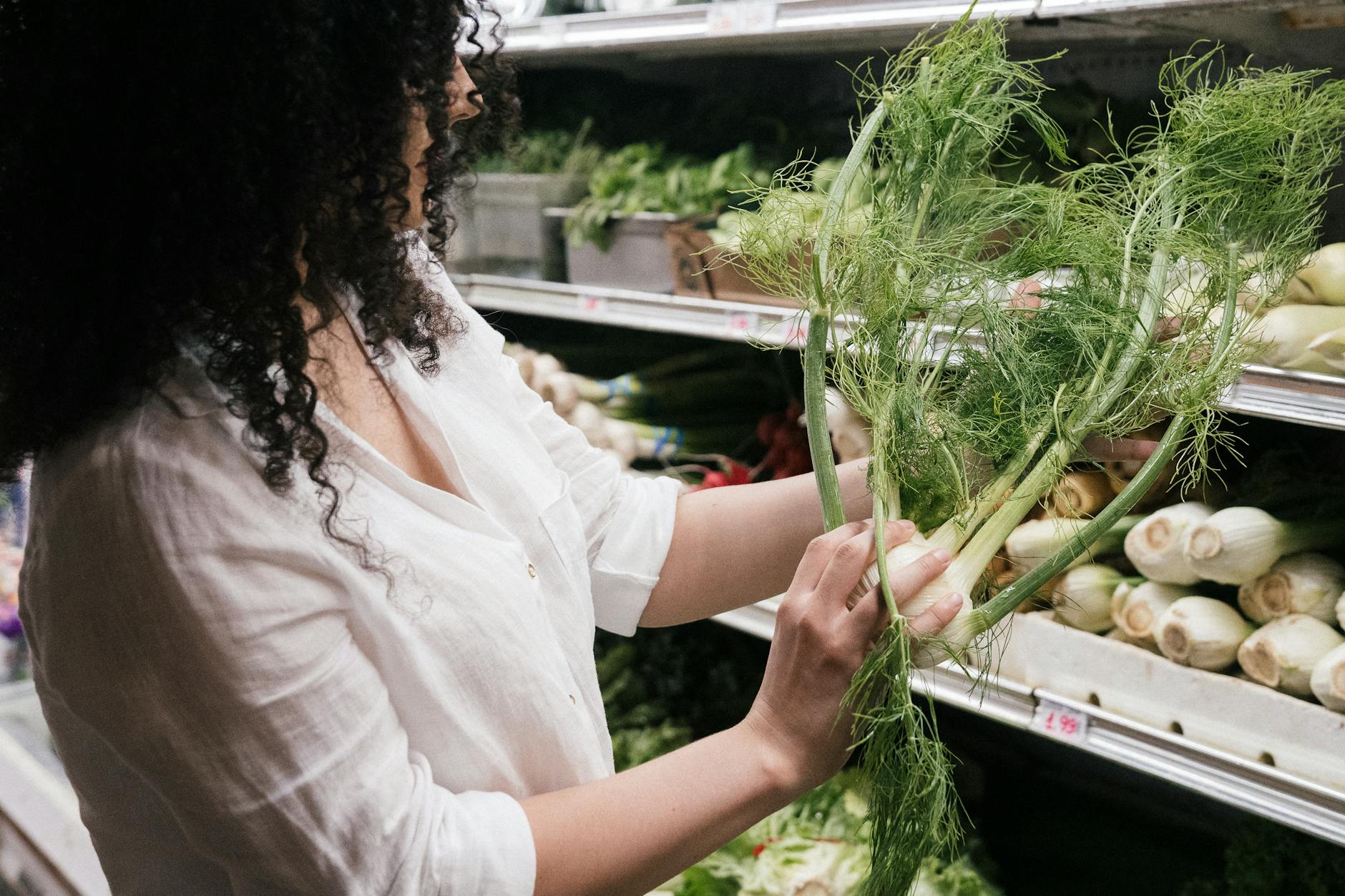 A woman in a white top choosing organic fennel in a grocery store produce section. - spring produce health