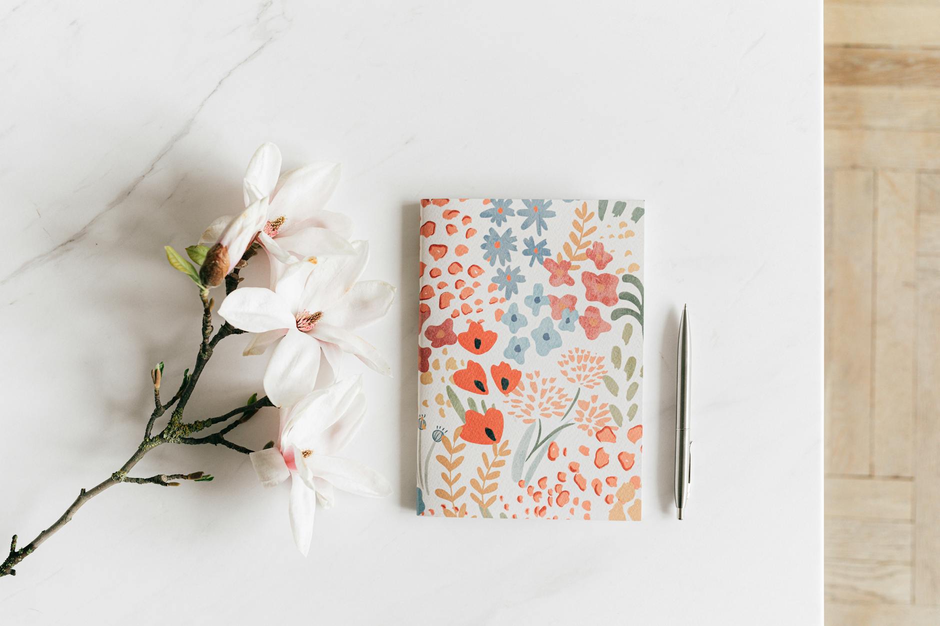 Top view composition of colorful floral notebook with silver pen arranged near twig of fragrant lush white Magnolia placed on white desk - spring reset journal prompts
