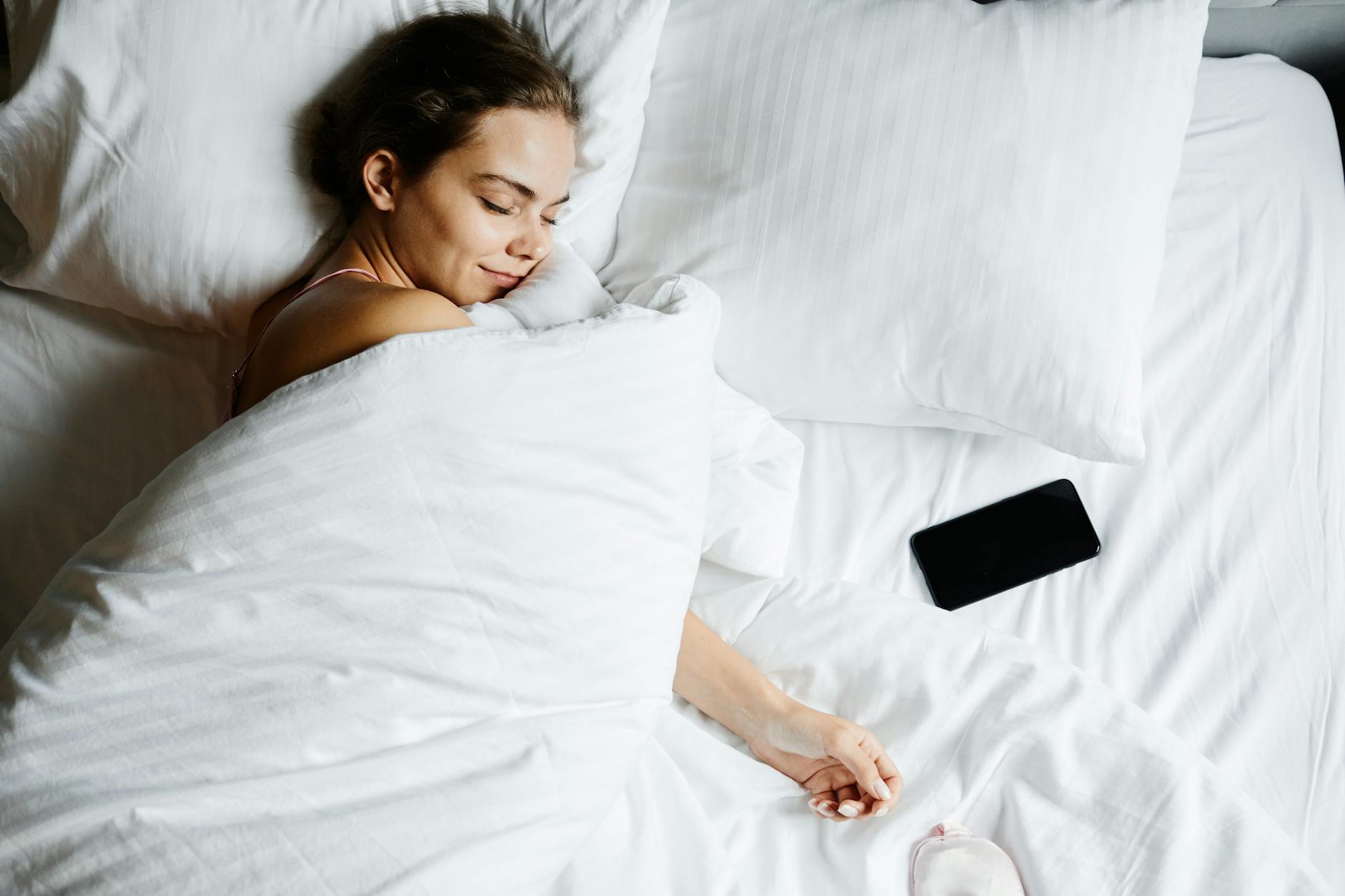 A young woman sleeps comfortably in bed with a smartphone beside her, wrapped in white sheets. - spring sleep reset