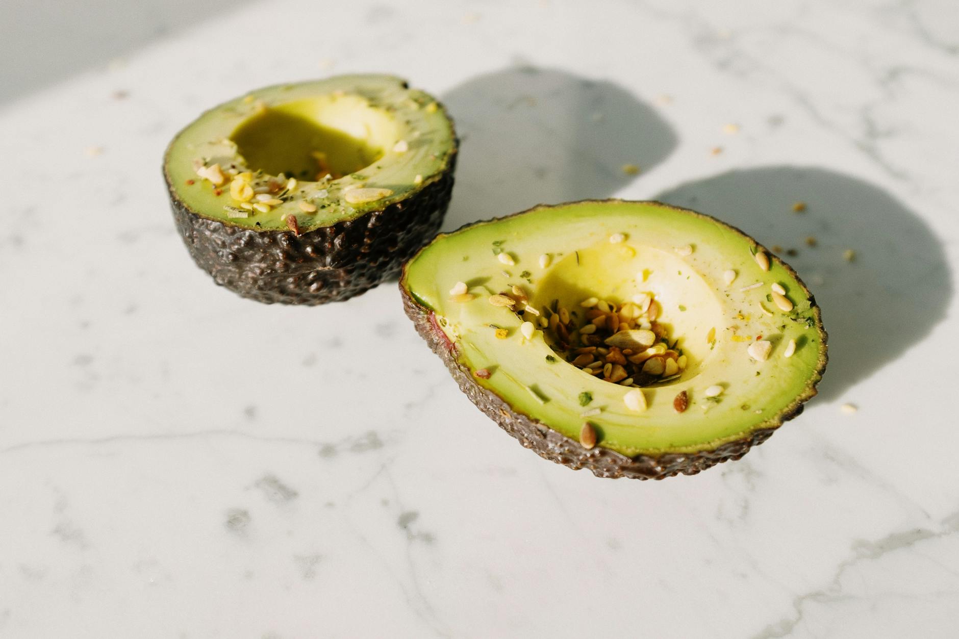 From above of green halved avocado without kernel sprinkled with various seeds placed on white marble desk served for healthy vegan breakfast preparation - spring superfoods skin