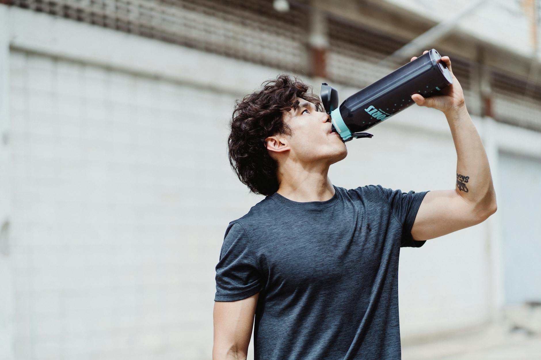 A young man in a casual shirt rehydrates with water from a bottle outdoors. - spring workout hydration