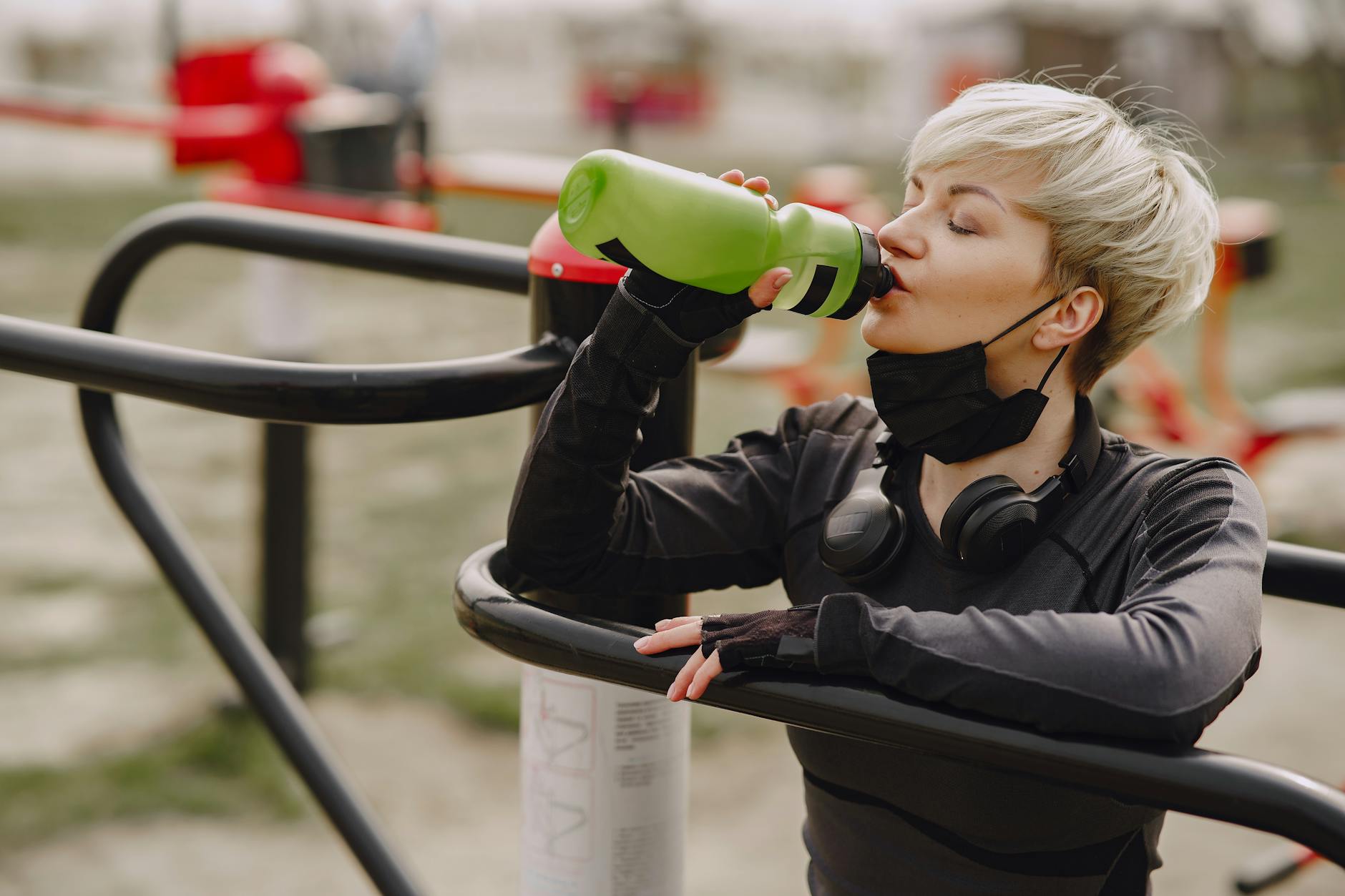 Calm female in sportswear and sport gloves with headphones on neck leaning on street exercise machine  drinking water from plastic bottle - staying hydrated spring
