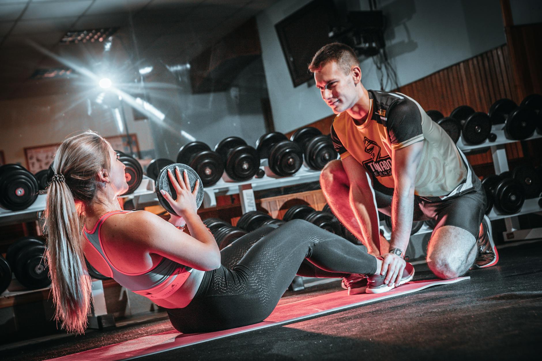 A personal trainer assists a woman doing sit-ups with a medicine ball in a gym setting. - advanced core training