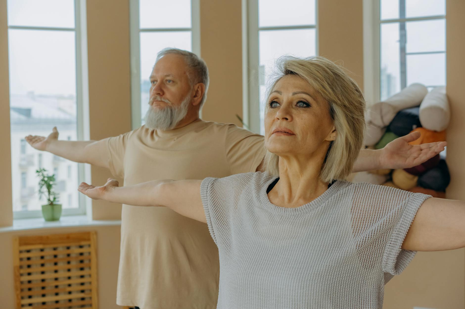 Elderly couple practicing yoga with arms outstretched in a bright indoor setting. - balance exercise social life
