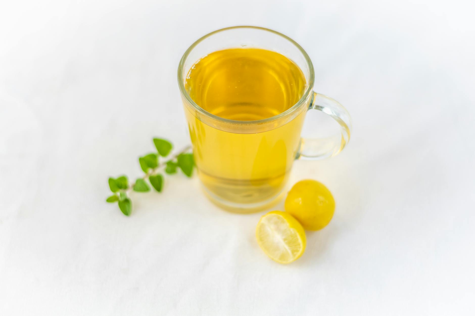A clear glass of lemon herbal tea accompanied by fresh herbs on a white surface, offering a refreshing drink. - boost immunity naturally