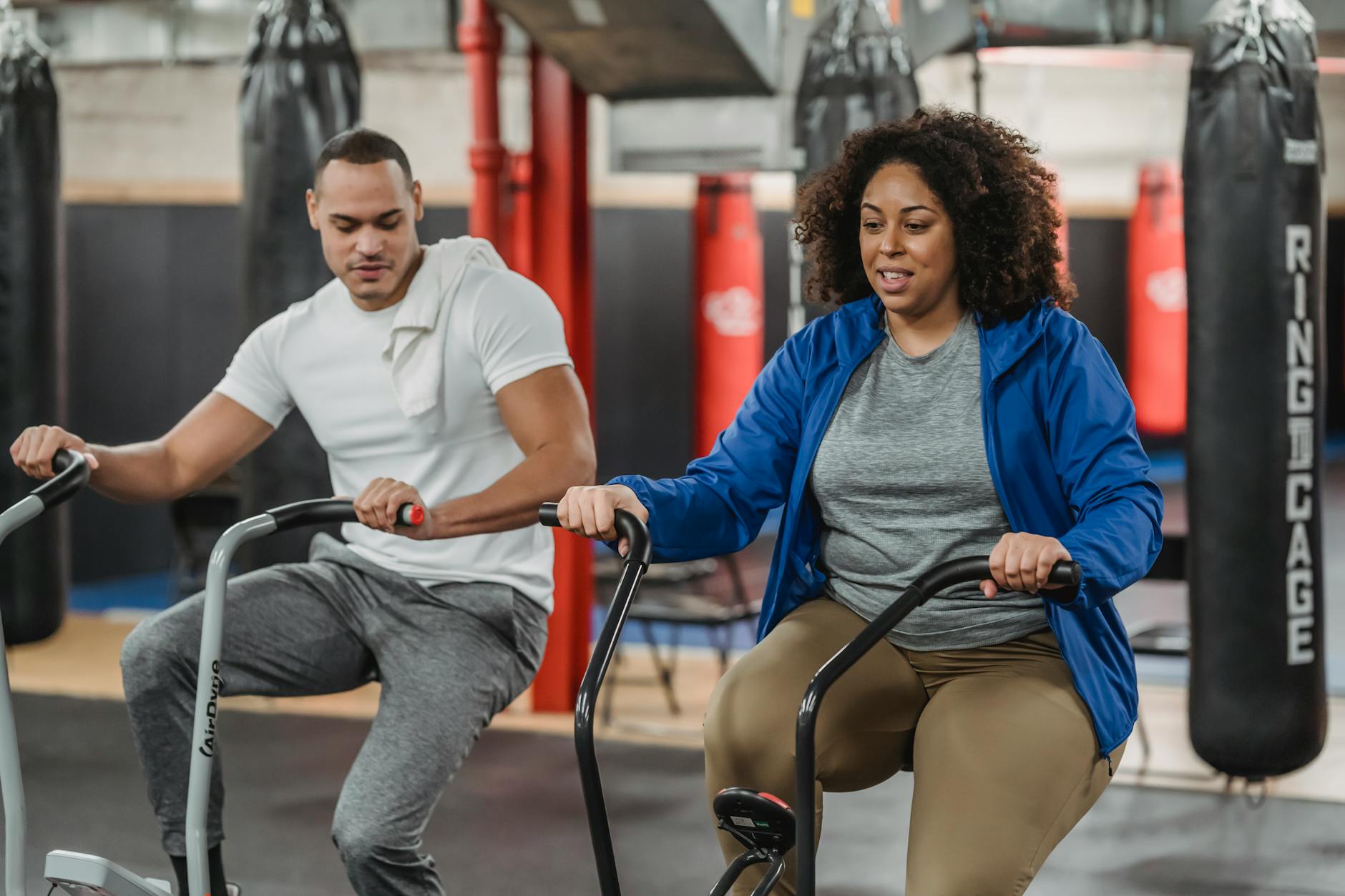 Plus sized African American female in sportswear exercising on stationary bicycle near fit muscular male coach in modern gym - cardio for weight loss