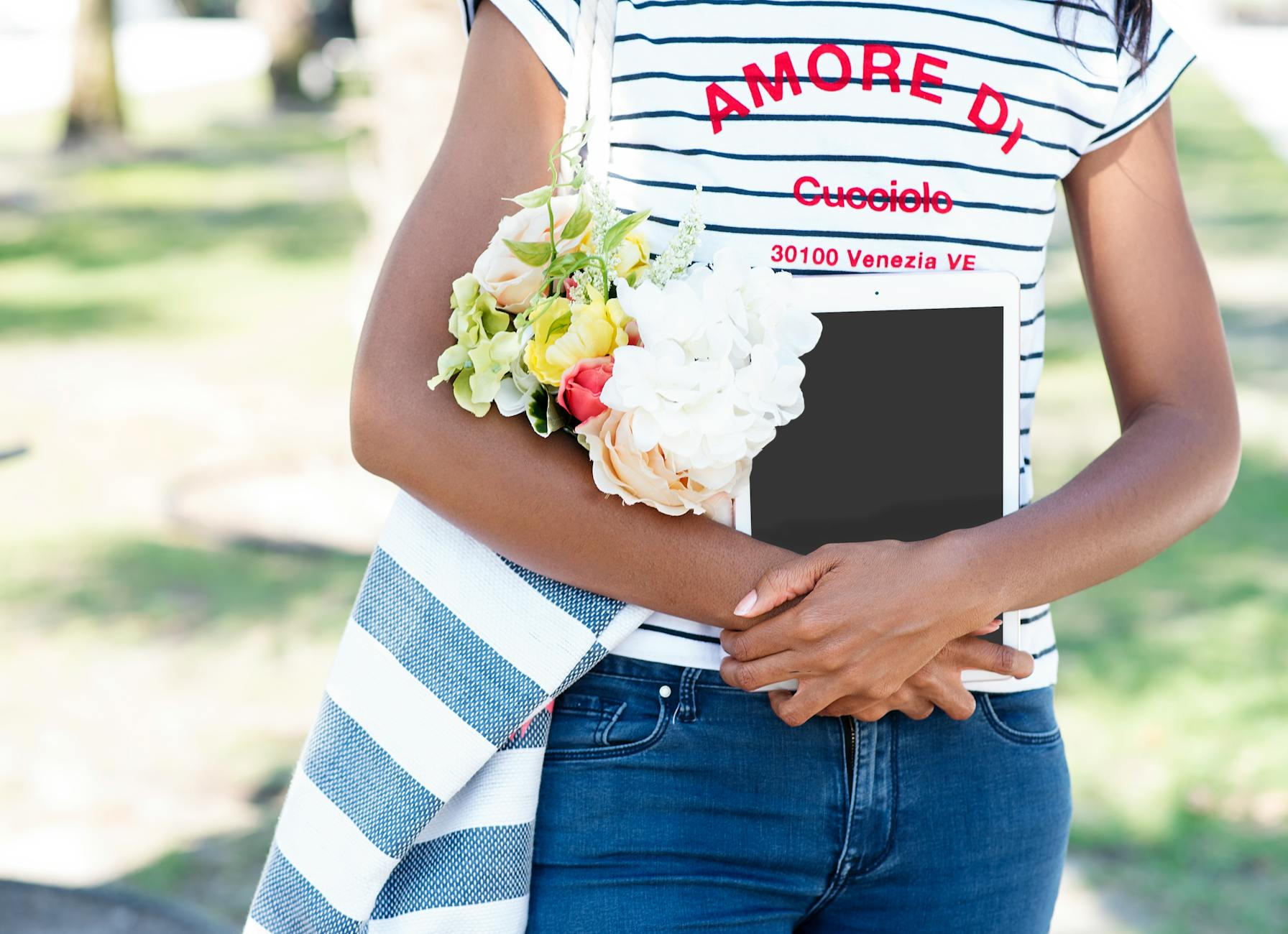 A young woman in a striped shirt holds a tablet and a bouquet of flowers in a sunny outdoor park. - digital declutter spring