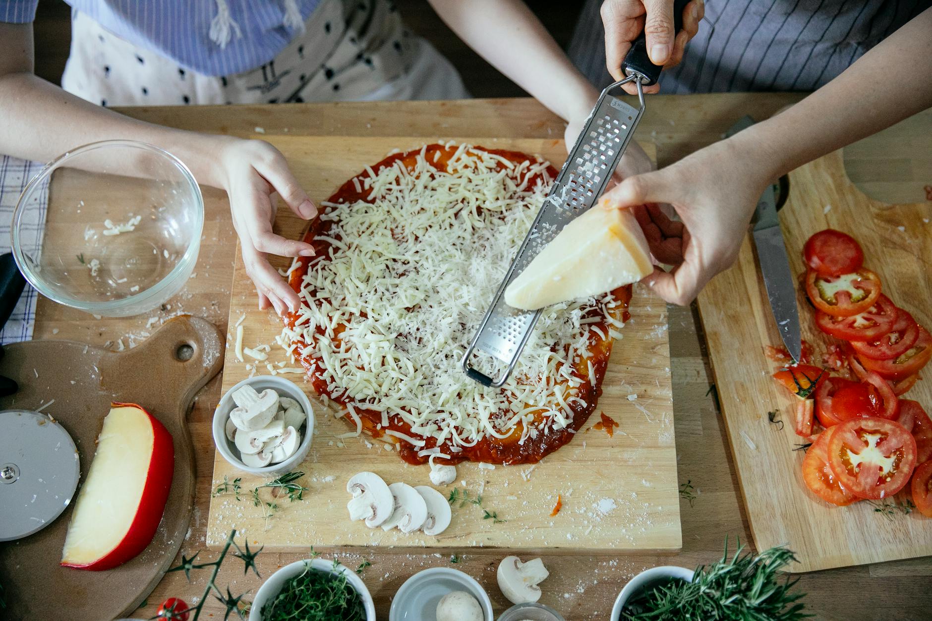Overhead crop women grating hard cheese together while cooking pizza on wooden table with various ingredients and utensil in kitchen - easy weeknight dinners