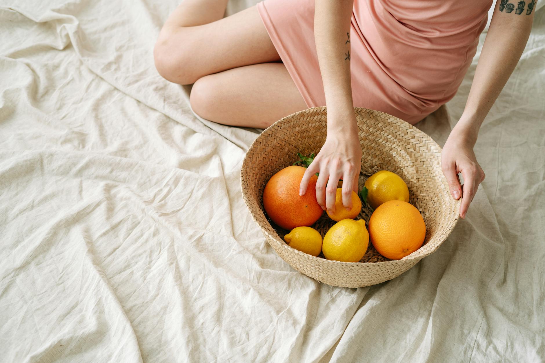 A woman in a pink dress holding a basket of fresh citrus fruits, including oranges and lemons. - glowing skin foods