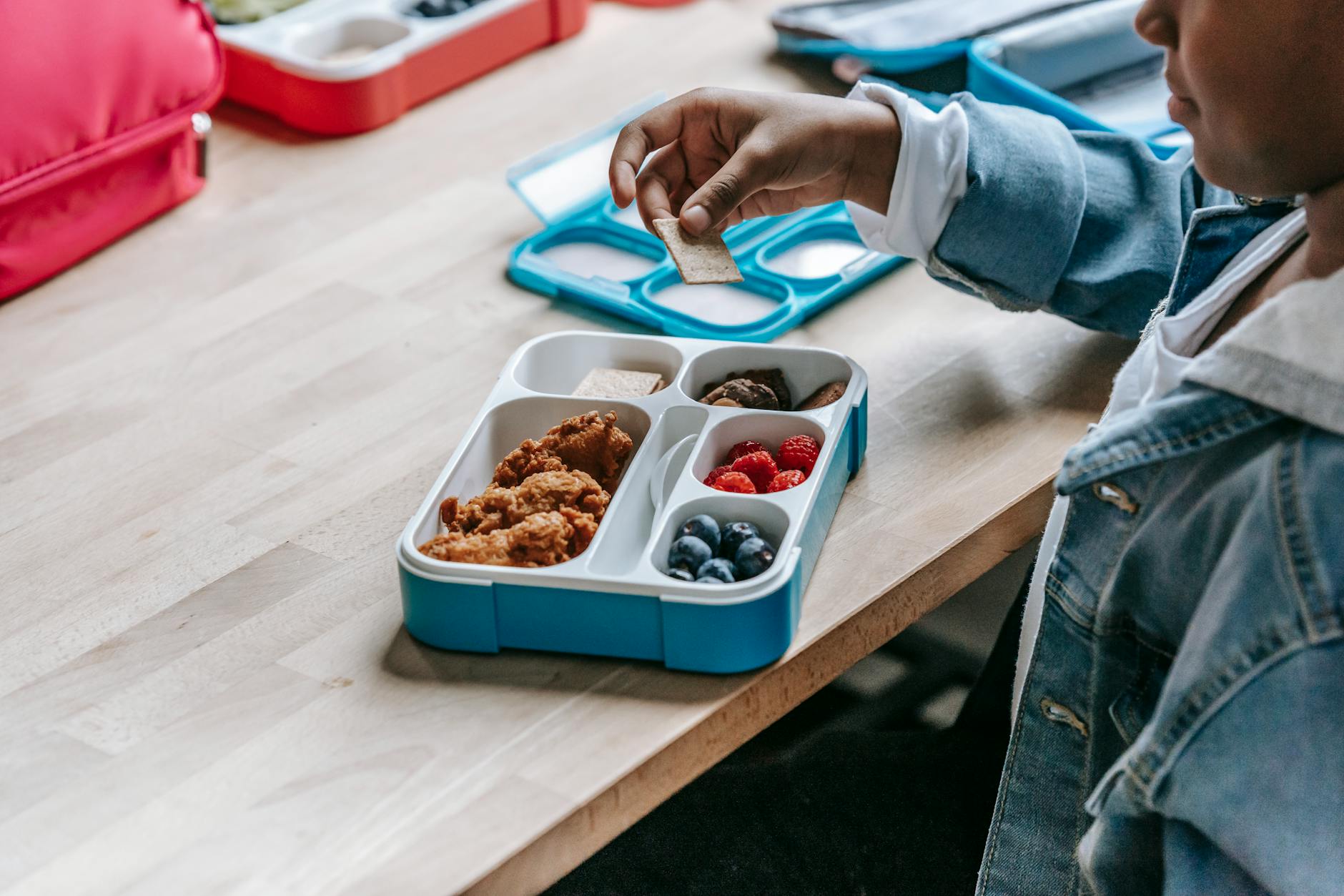 From above side view of crop unrecognizable ethnic schoolkid at table with lunch container full of tasty food - healthy lunch prep