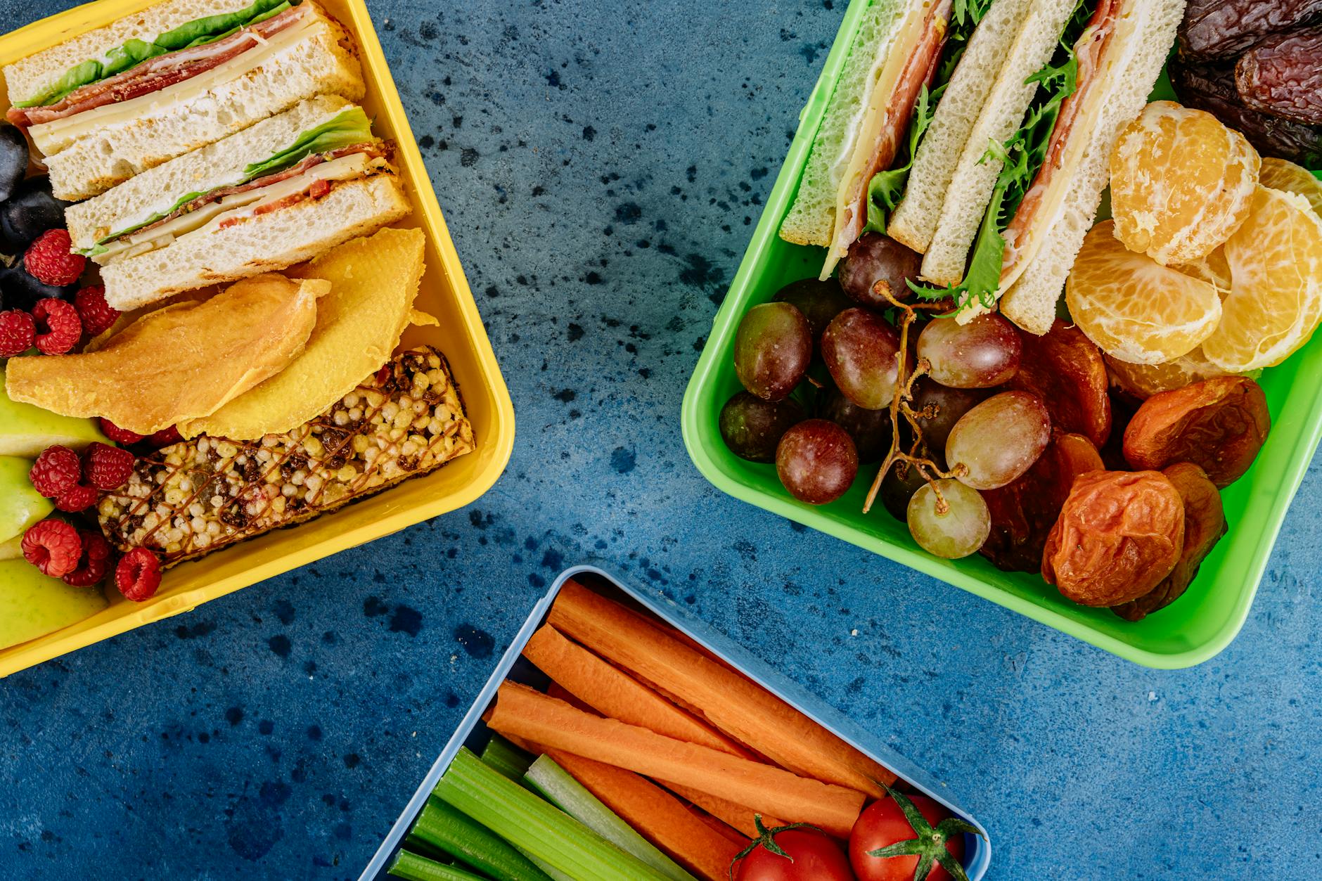 Top view of vibrant lunch boxes filled with sandwiches, fruits, and vegetables on a blue surface. - healthy lunch prep