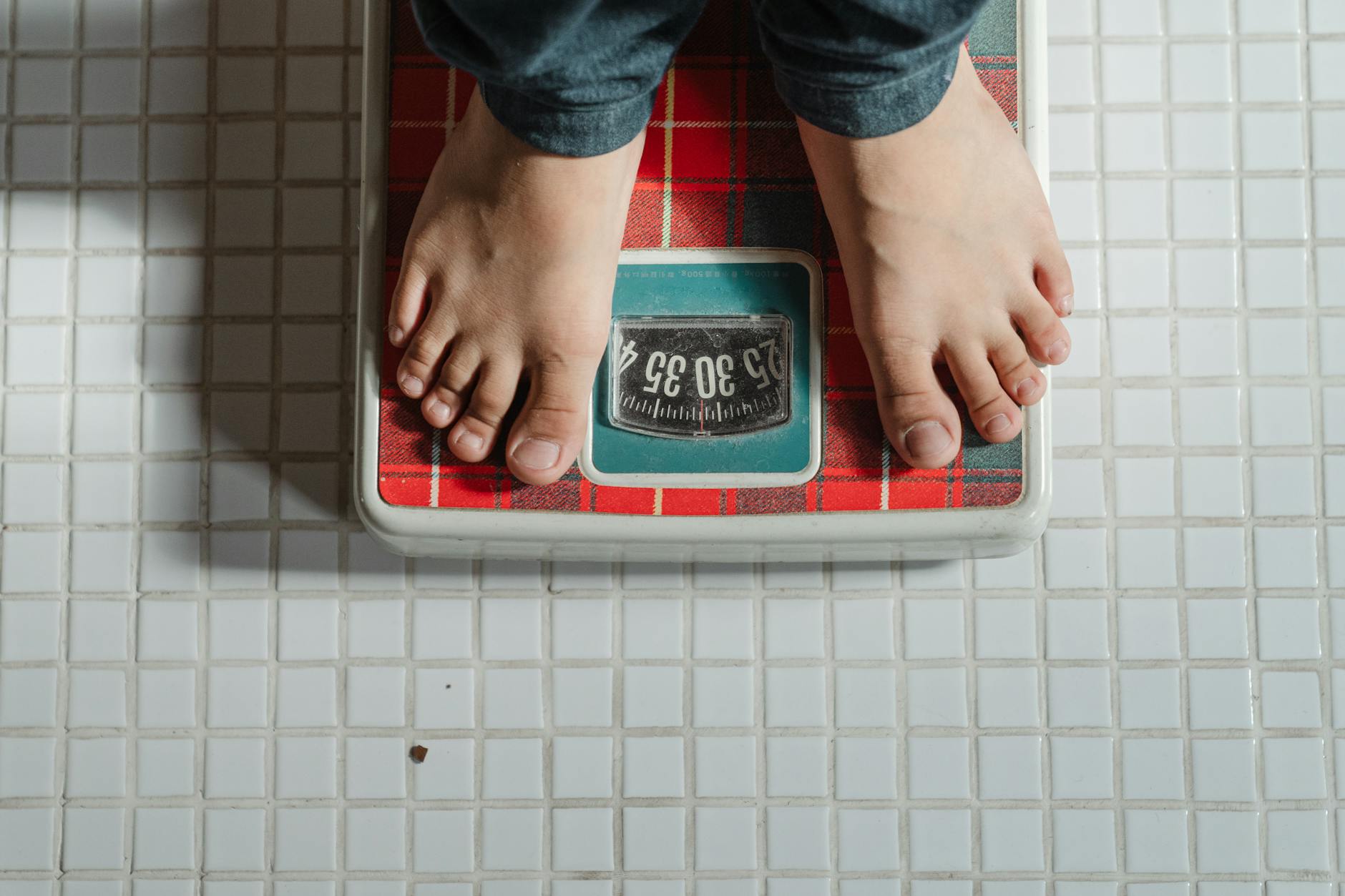 From above crop anonymous barefoot child in jeans standing on weigh scales on tiled floor of bathroom - healthy weight loss