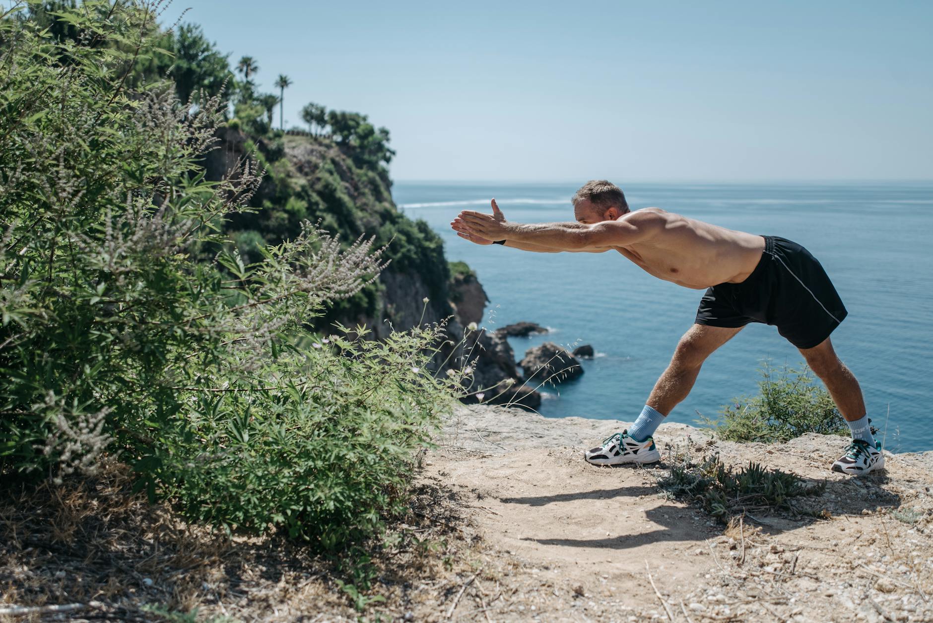 A shirtless man performs stretching exercises on a scenic cliff overlooking the ocean. - hiking endurance training