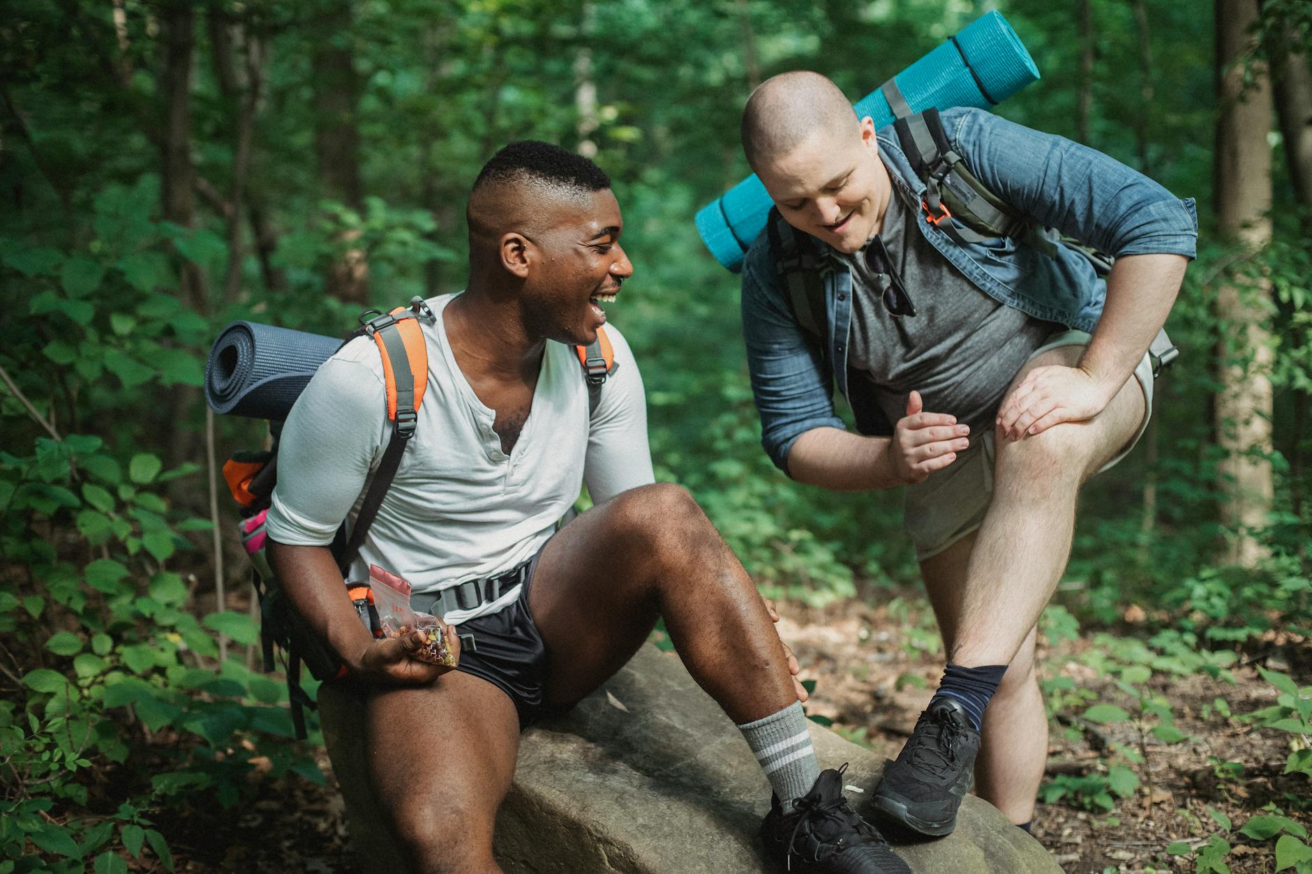 Happy men in activewear with backpacks laughing while having break during hiking in lush forest - hiking energy snacks
