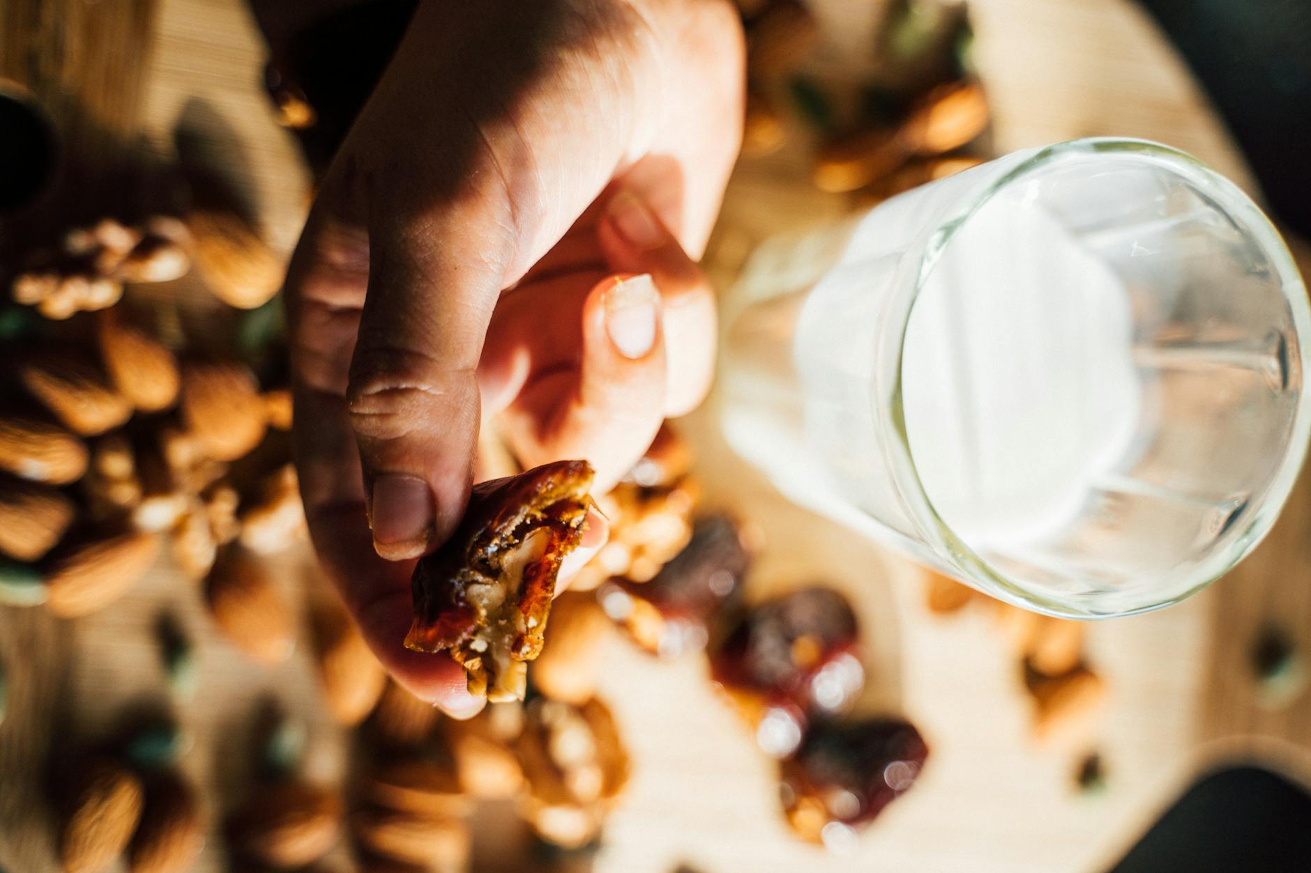 Close-up of a hand holding a date with nuts, surrounded by almonds and walnuts, next to a glass of milk. - hiking energy snacks