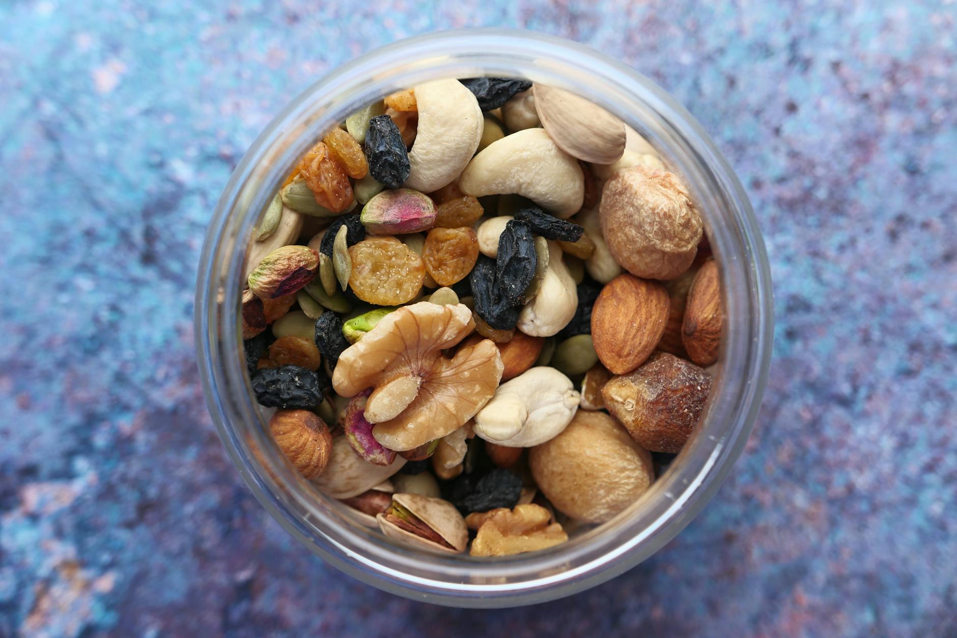 Top view of a variety of nuts and dried fruits in a jar against a blue background. - hiking trail snacks