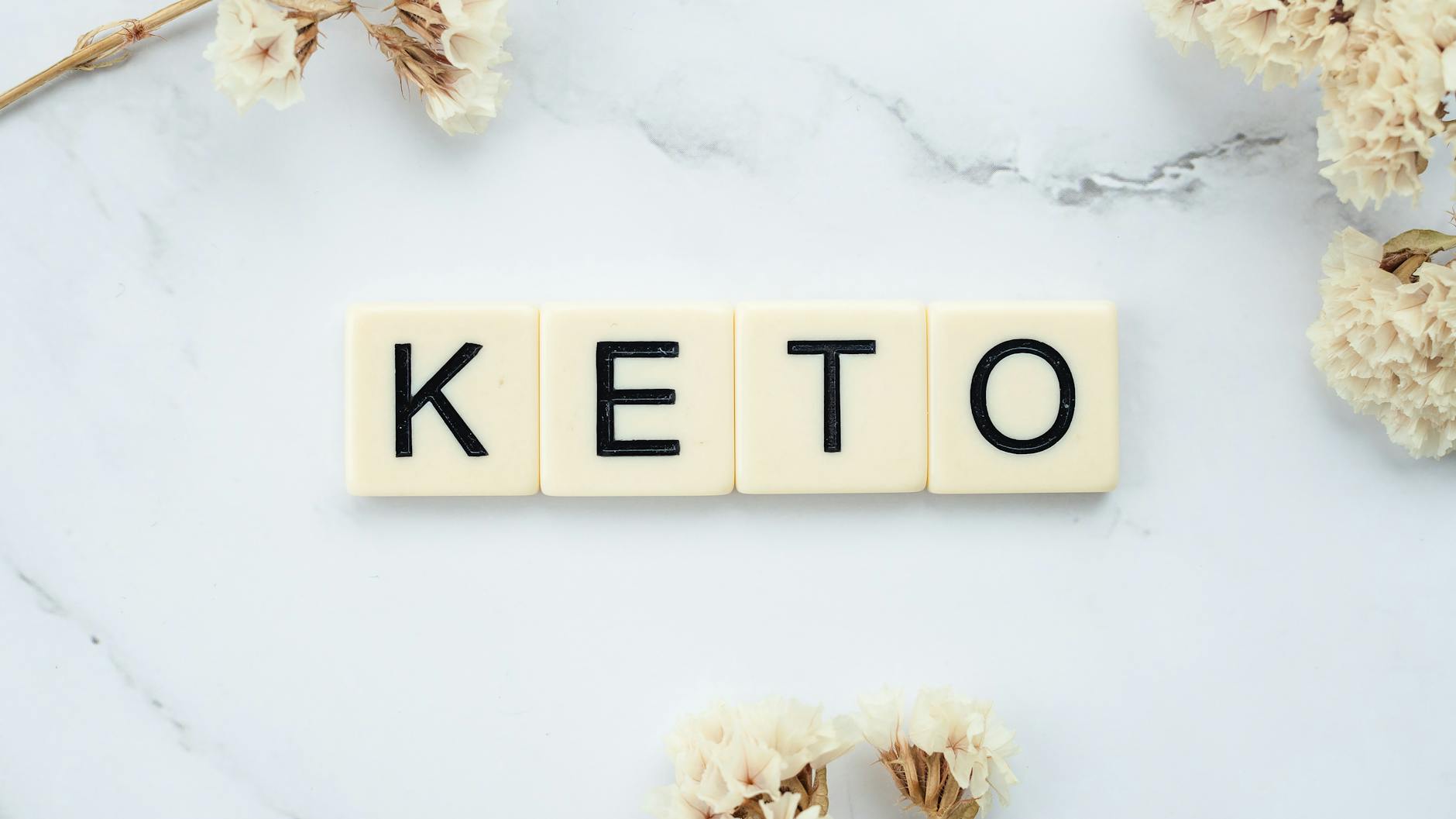 Scrabble tiles spelling 'Keto' on a marble surface with white flowers, symbolizing a ketogenic lifestyle. - low fat low carb