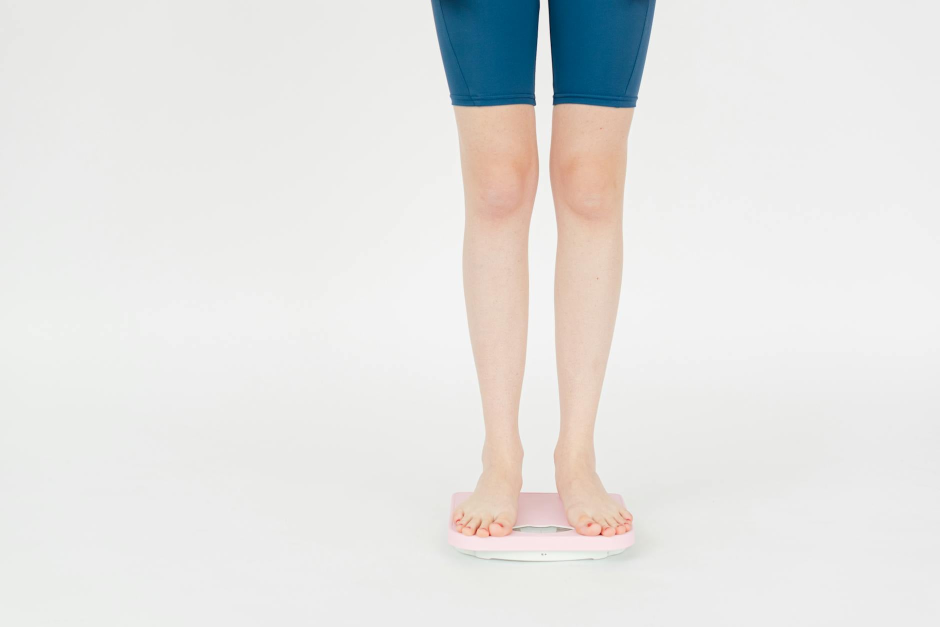 Crop anonymous female in sportswear measuring weight on scales for body control against white background - non-scale victories